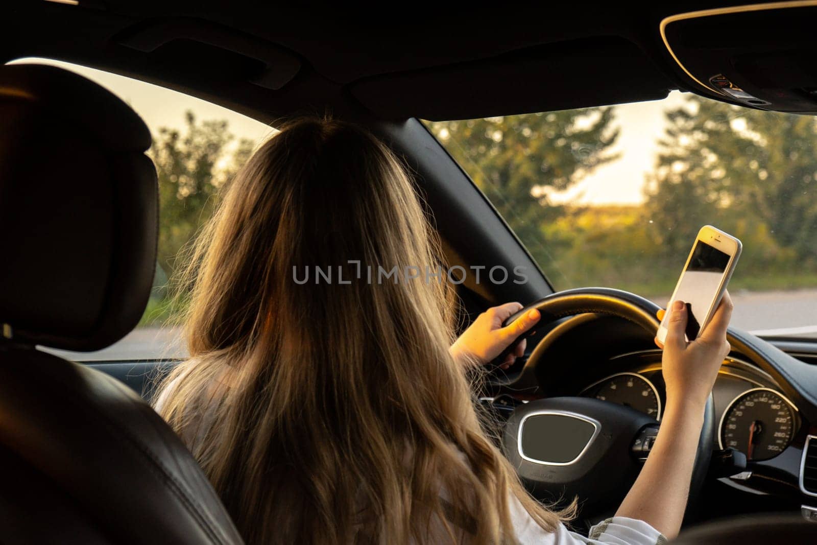 Young woman using mobile phone while driving car on highway road during sunset. Womandriver has accident calling with smartphone for help. Business woman busy driving by anna_stasiia