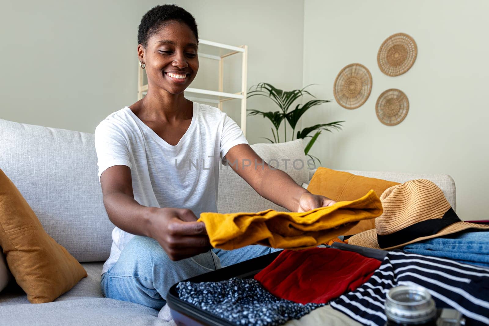 African American young woman packing suitcase for summer vacation trip sitting on the sofa. Holiday, tourist, vacation concept.