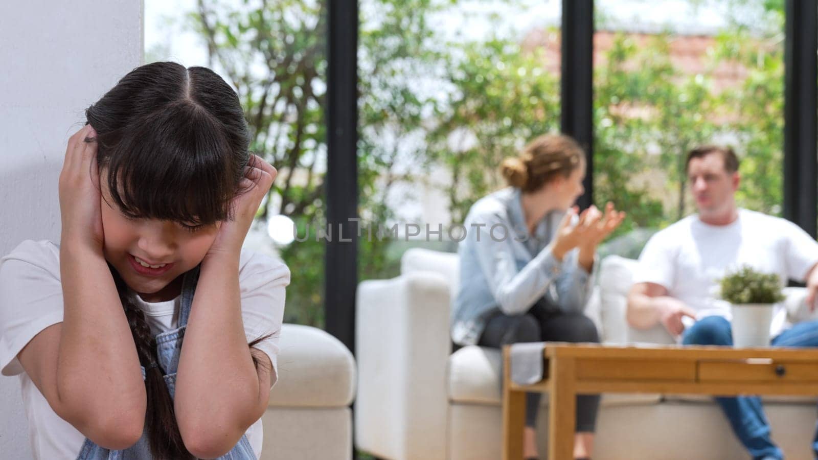 Stressed and unhappy young girl huddle in corner, cover her ears blocking sound of her parent arguing in background. Domestic violence at home and traumatic childhood develop to depression. Synchronos