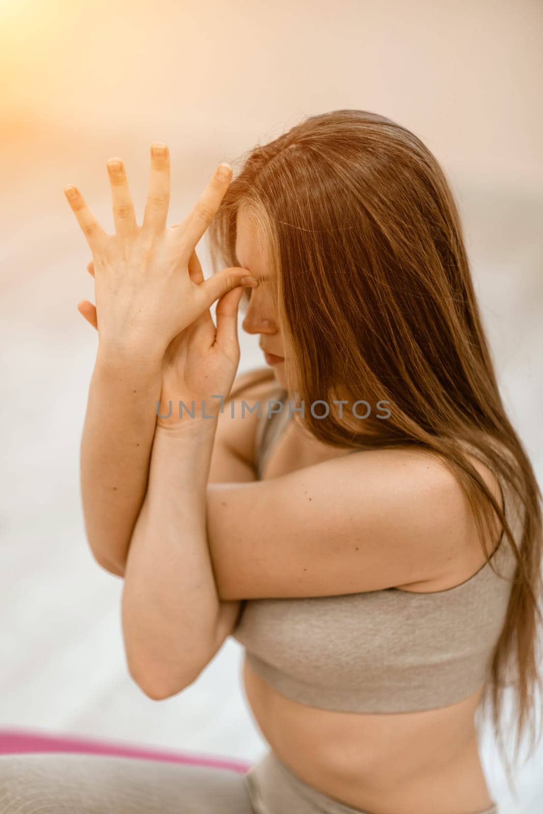 Young woman doing yoga in the gym. A girl with long hair and in a beige tracksuit stands in a cow pose on a pink carpet. A woman performs Gomukhasana. by Matiunina