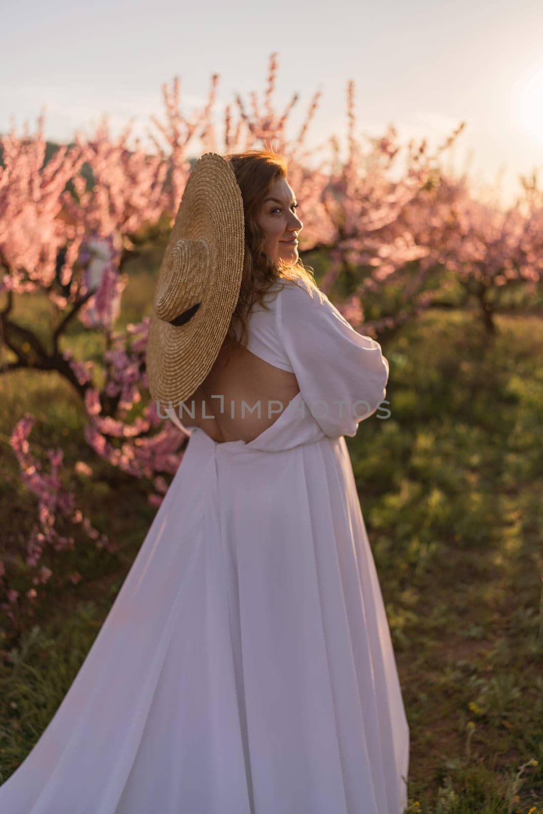 Woman blooming peach orchard. Against the backdrop of a picturesque peach orchard, a woman in a long white dress and hat enjoys a peaceful walk in the park, surrounded by the beauty of nature