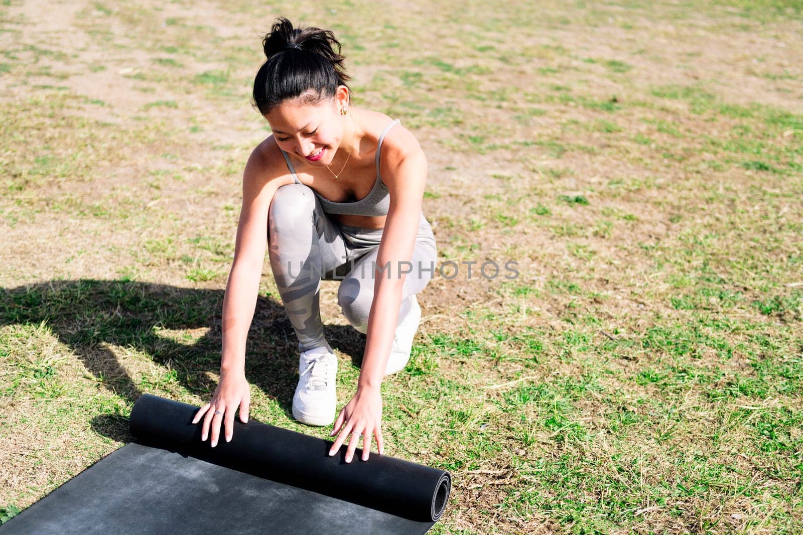 smiling young asian woman in sportswear rolling her mat for a yoga session on the grass in the park, healthy and active lifestyle concept, copy space for text