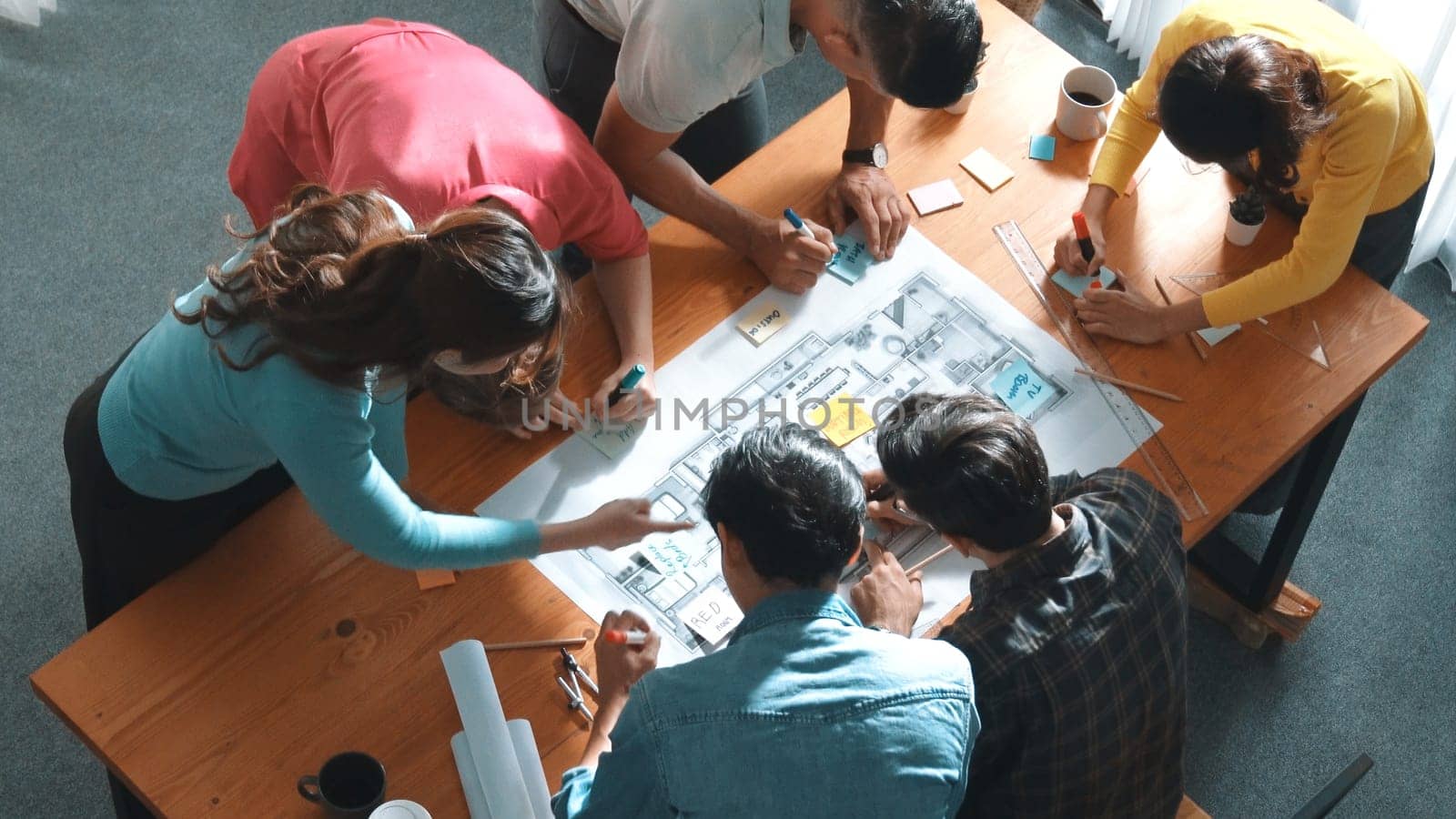 Top view of smart civil engineer working and writing blueprint while sitting at meeting table with blueprint, architectural equipment and colorful sticky note. Diverse team joining meeting. Symposium.
