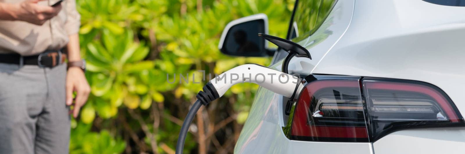 Young man use smartphone to pay for electricity at public EV car charging station green city park. Modern environmental and sustainable urban lifestyle with EV vehicle. Panorama Expedient