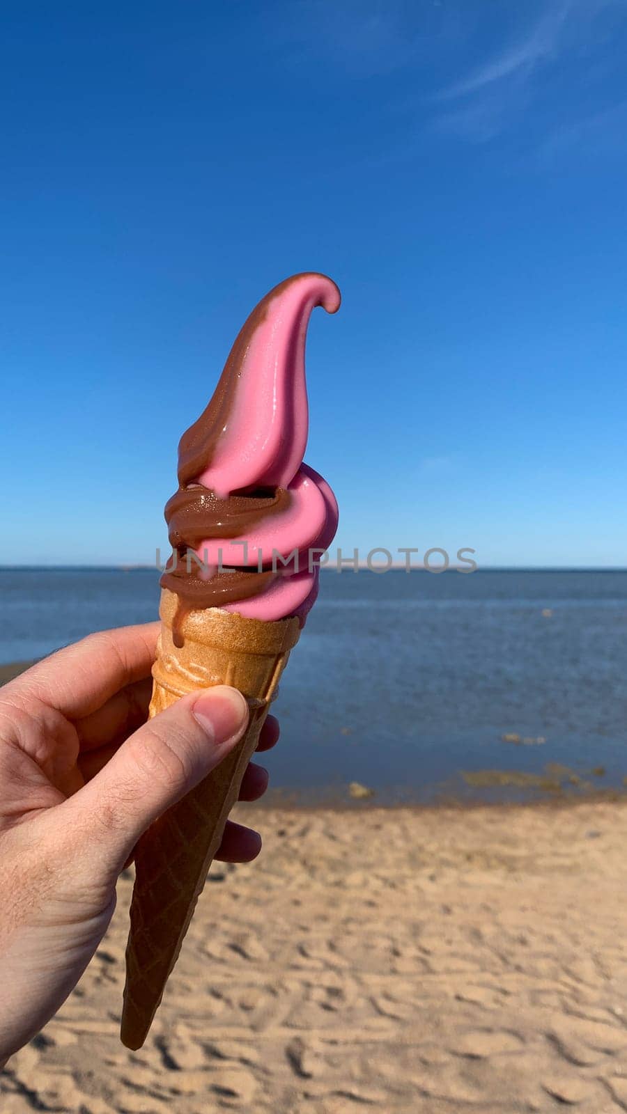 Ice cream in cone melting in sun in hand against backdrop of beach by the sea, vertical photo of travel concept