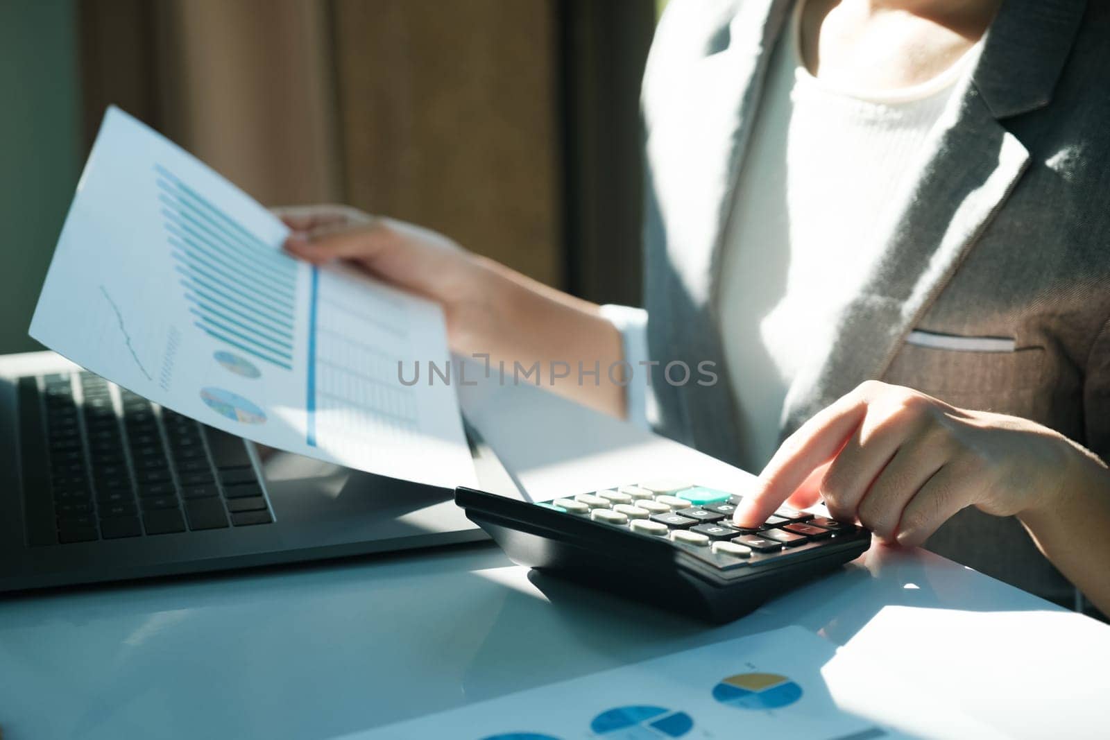 A woman is sitting at a desk with a calculator and a piece of paper with graphs on it. She is focused on the paper and the calculator, possibly working on a project or analyzing data
