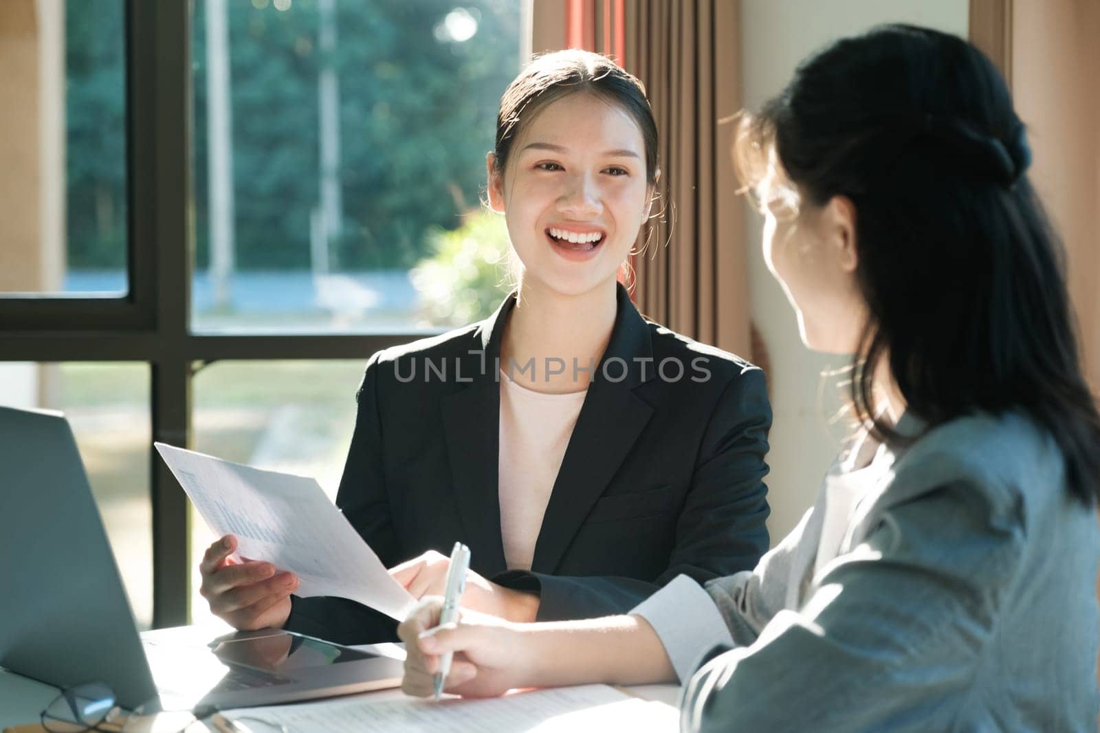 Two women in business suits are talking to each other. One of them is holding a piece of paper