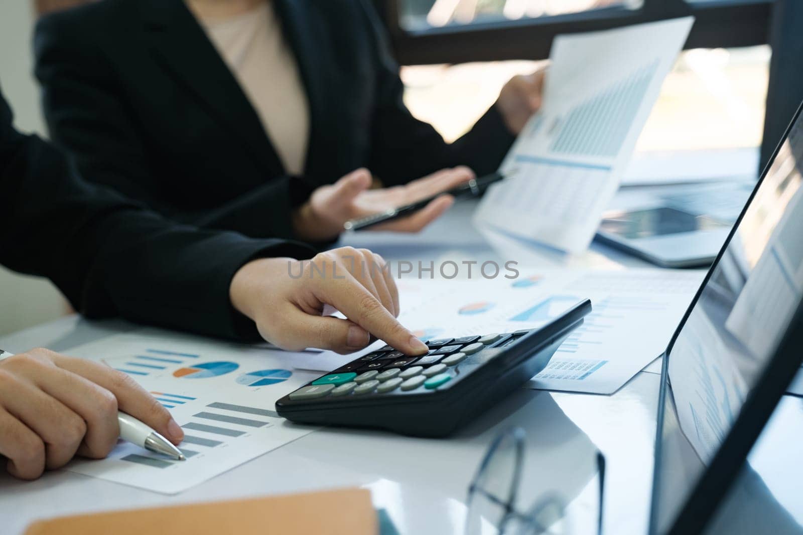 Two women are working on a spreadsheet, one of them pointing at a calculator. Scene is focused and professional