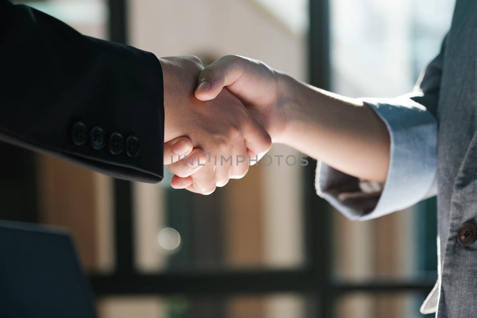 Two people shaking hands in a business setting. The man is wearing a suit and tie, while the woman is wearing a blazer. The handshake is a sign of agreement or partnership between the two individuals