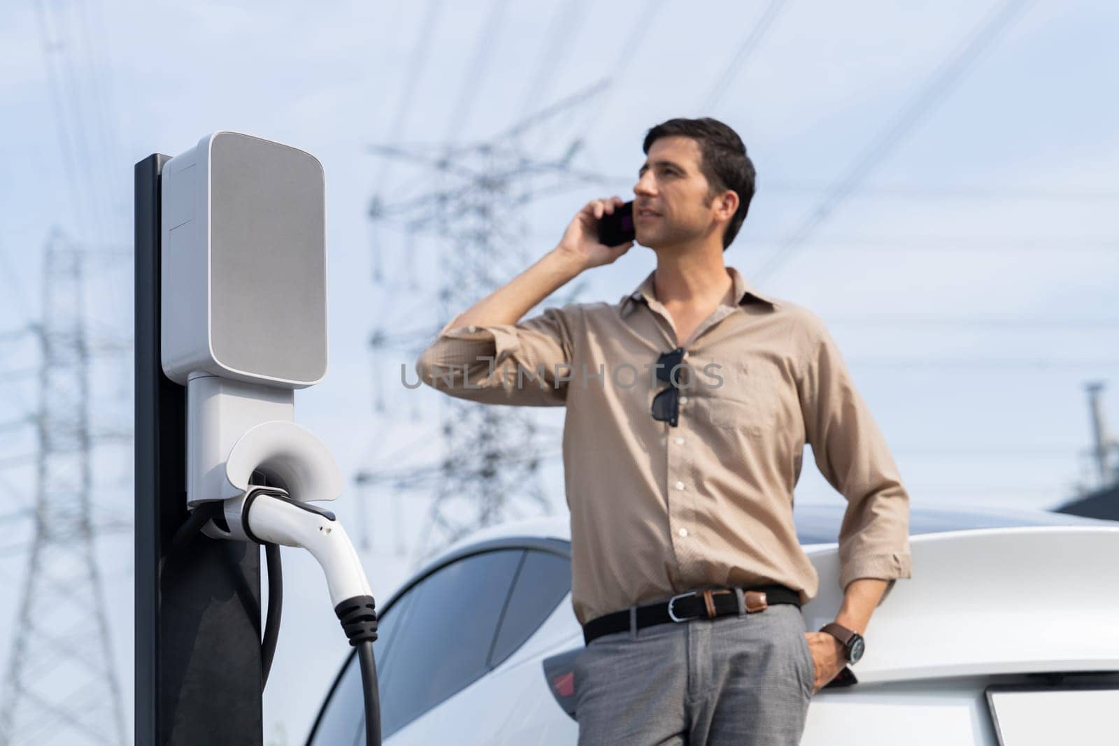 Man talking on the phone while recharge EV car battery at charging station connected to power grid tower electrical as electrical industry for eco friendly car utilization.Expedient