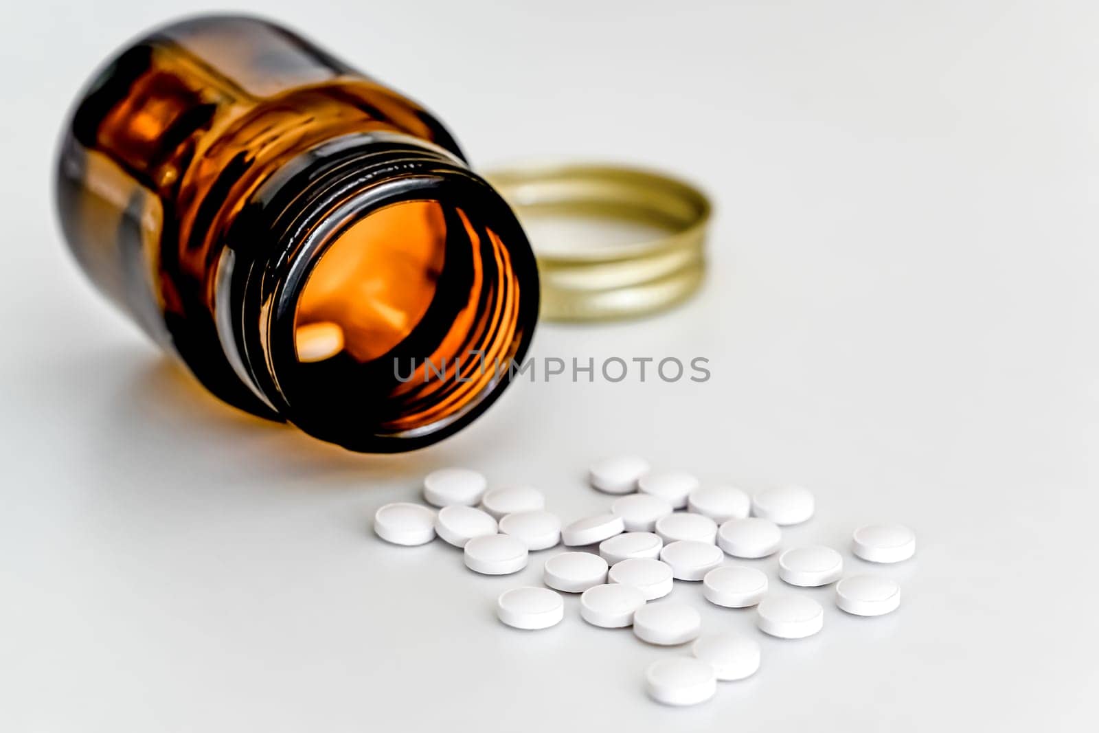 White Pills Scattered on Table Next to Brown Glass Vial, on White Background, Close-up