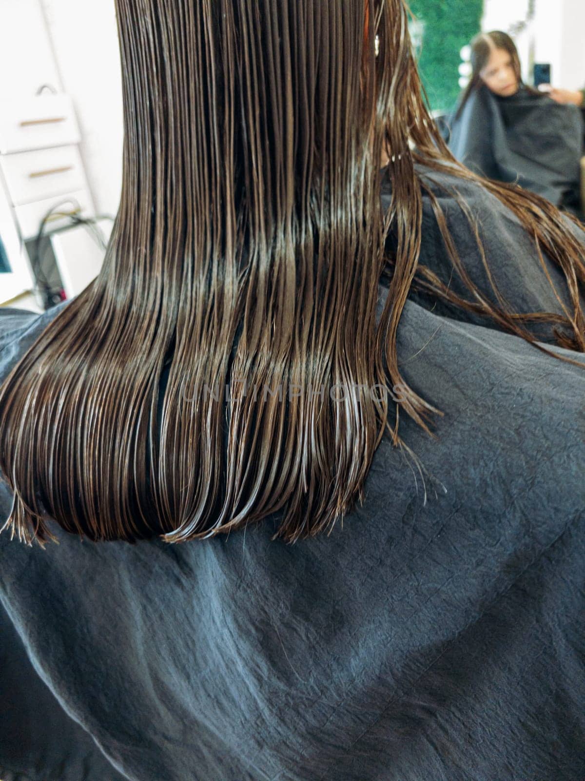 In the bustling atmosphere of a salon, a little girl sits patiently as a stylist with polished red nails carefully trims her damp hair. The sheen of the scissors and the precise hand movements capture the delicate process of styling a child's hair.
