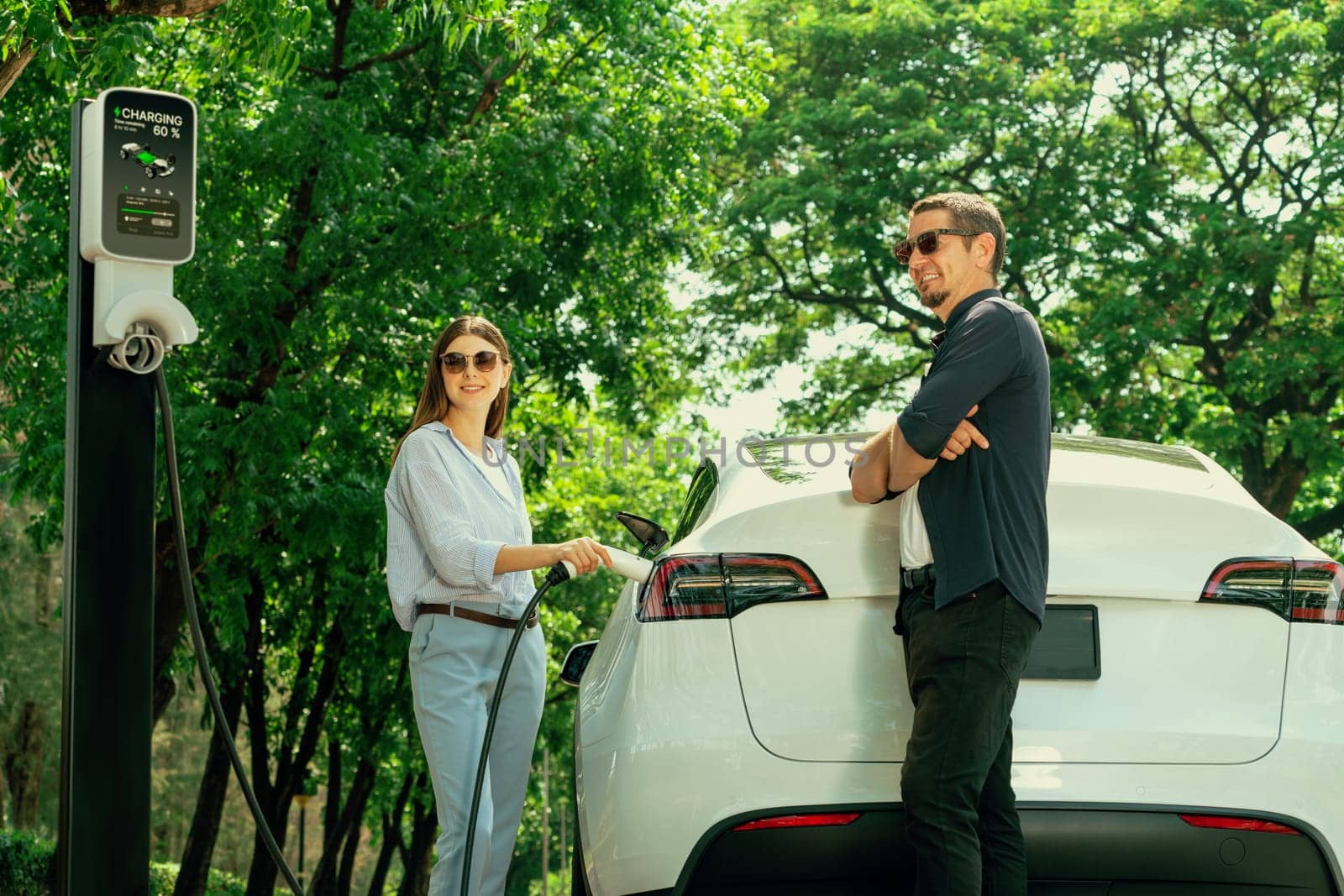 Lovely young couple wearing sun glasses recharging battery for electric car during road trip travel EV car in natural forest or national park. Eco friendly travel during vacation and holiday. Exalt