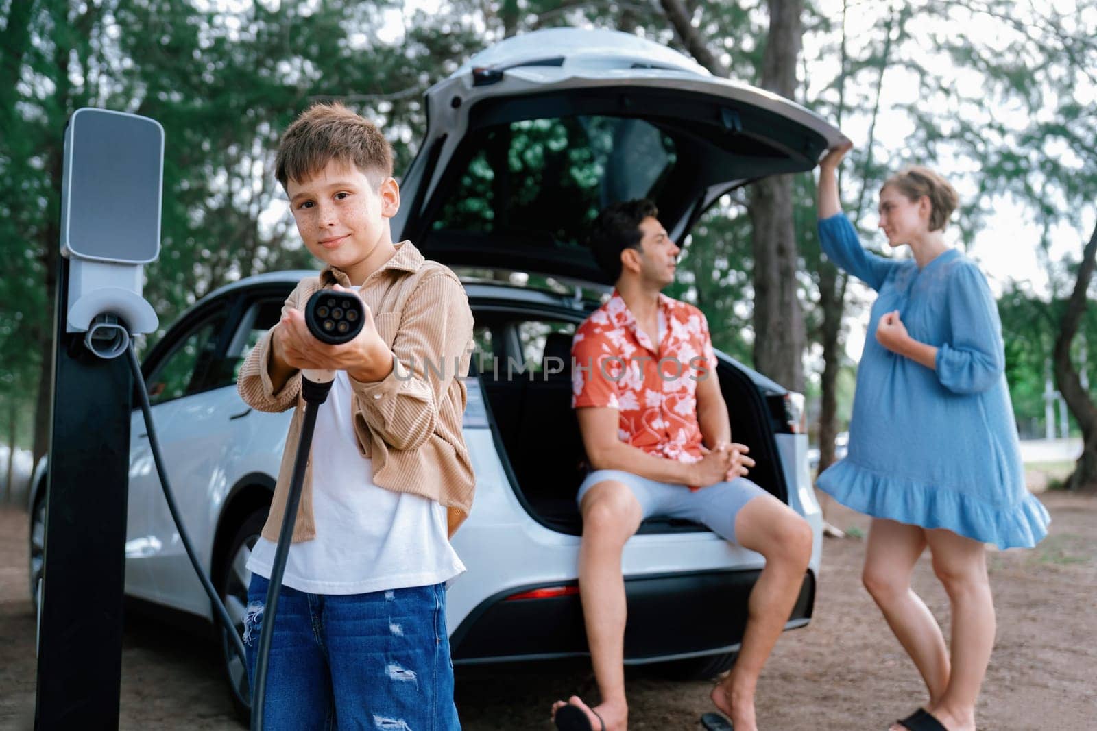 Little boy holding EV charger and point at camera with his family sitting on the trunk in background. Road trip travel with alternative energy charging station for eco-friendly car concept. Perpetual