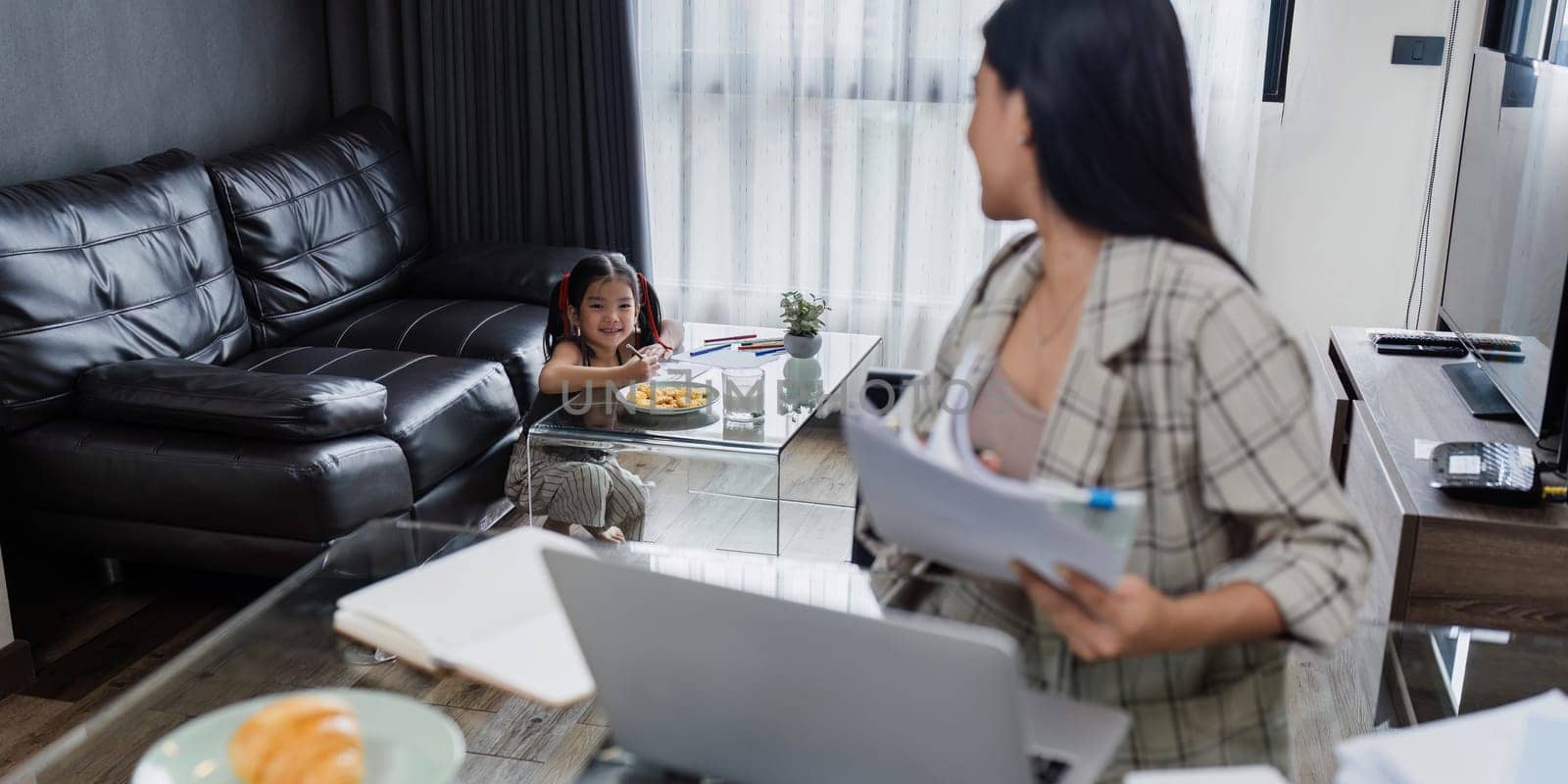 Business woman working from home and taking care of child while working, child doing activities in the background.
