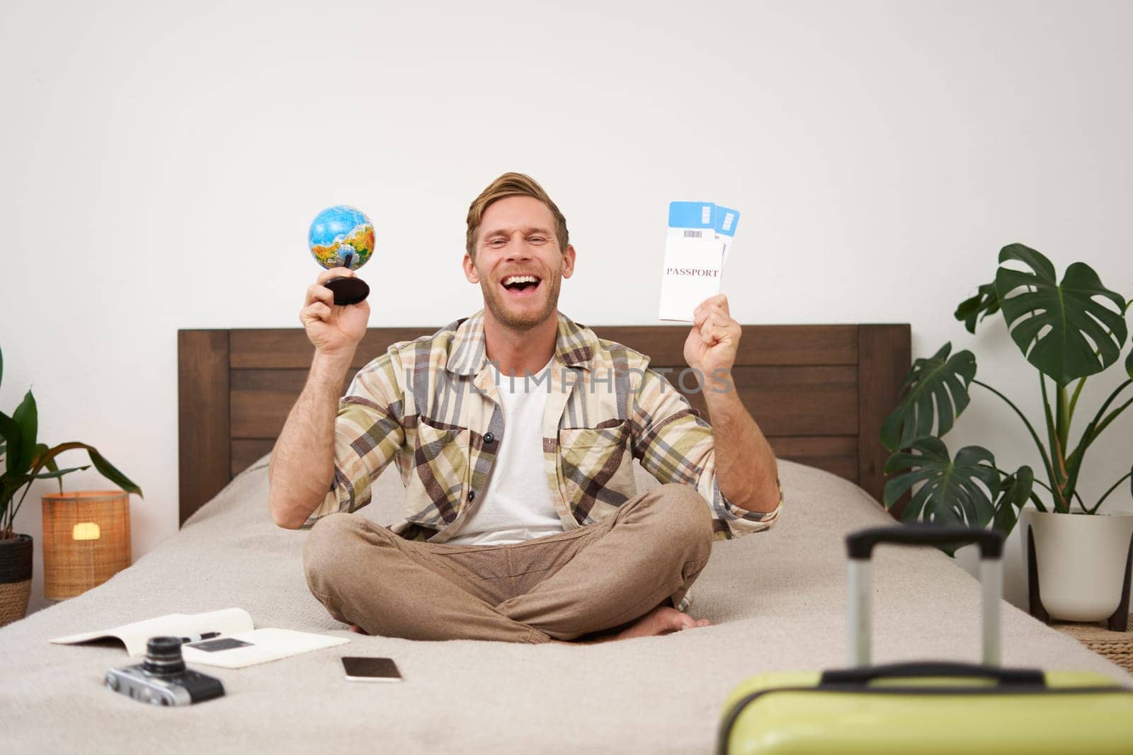 Portrait of cheerful, happy tourist, man sitting with plane tickets and a globe, packed suitcase for travelling around the world, going on vacation, excited about his holiday.