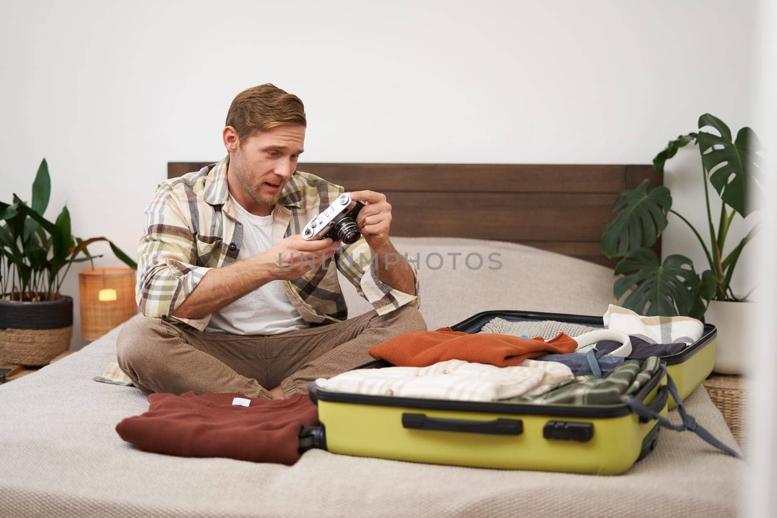 Portrait of young man looking at his digital camera screen, checking photo album on gadget, unpacking suitcase after holiday, travelling with luggage, sitting in hostel on a bed.