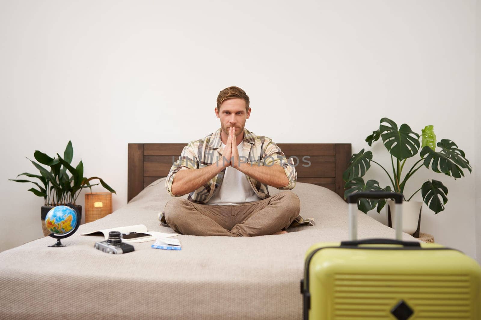 Portrait of traveller practice yoga, young man meditating before going on vacation, sitting with suitcase, globe and camera on bed in bedroom, relaxing, looking for peaceful holiday.