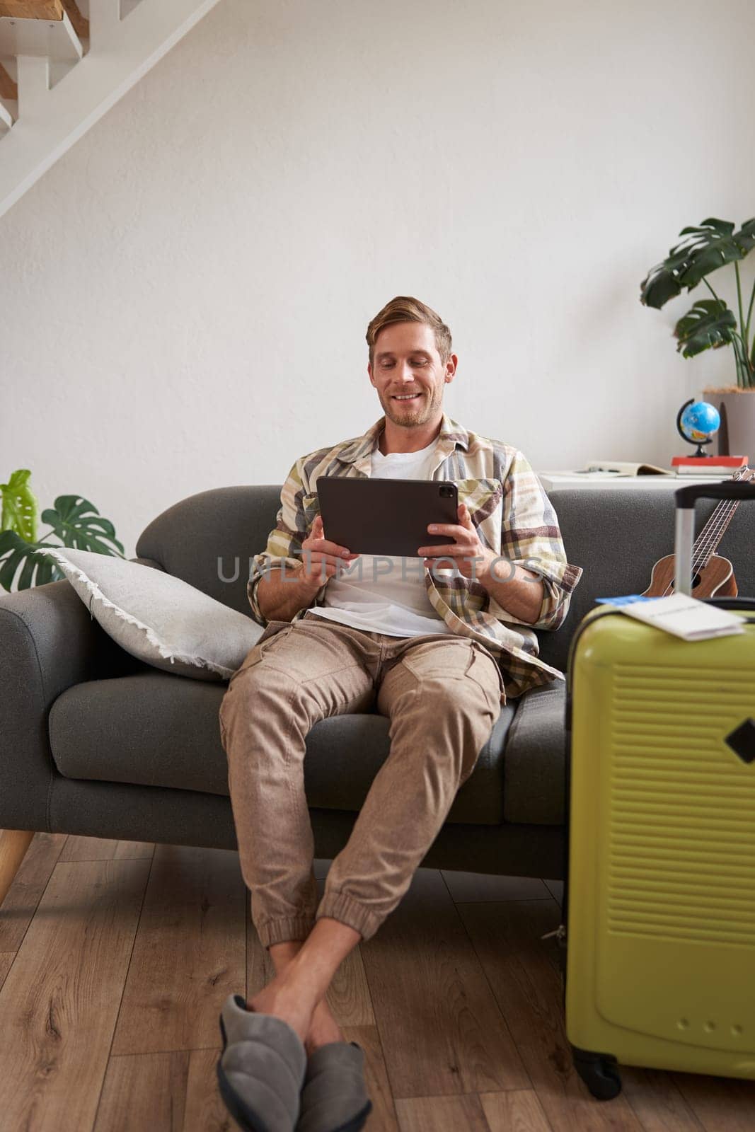 Vertical shot of man, tourist with suitcase, looking at digital tablet, reading about travel destination, booking hotel on gadget. Tourism concept