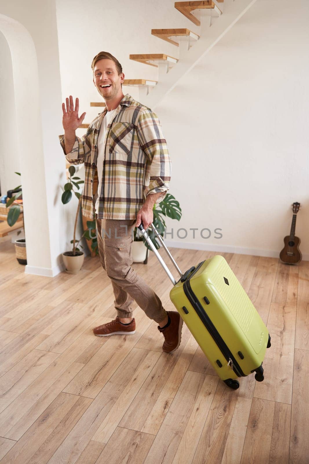 Full length photo of happy young man, tourist going on holiday, traveller leaving apartment with suitcase, waving hand at camera to say goodbye. Copy space