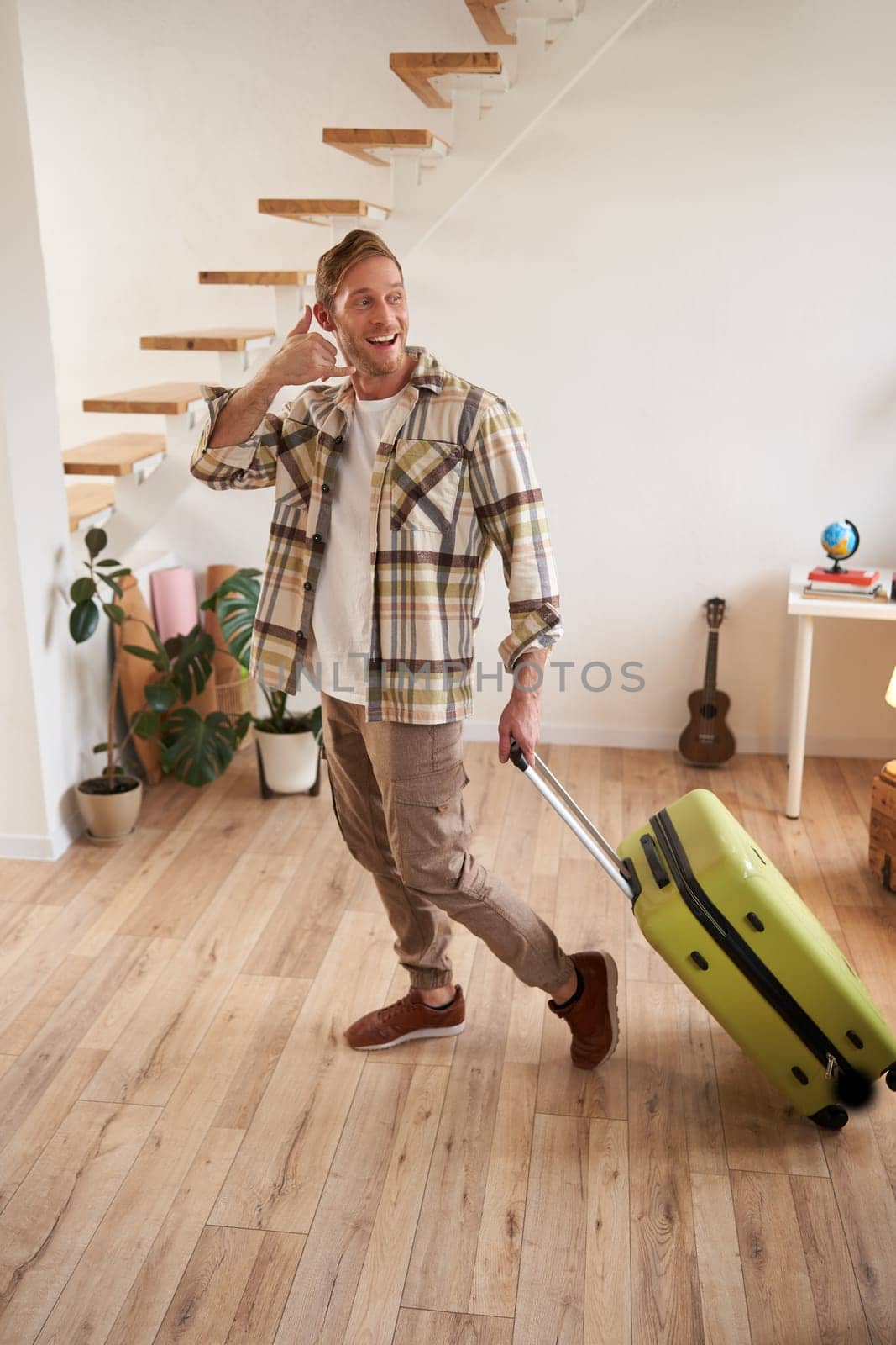 Portrait of handsome young man going on a trip, shows mobile phone hand sign gesture, going on vacation, leaving the apartment with suitcase, carrying luggage in hand.