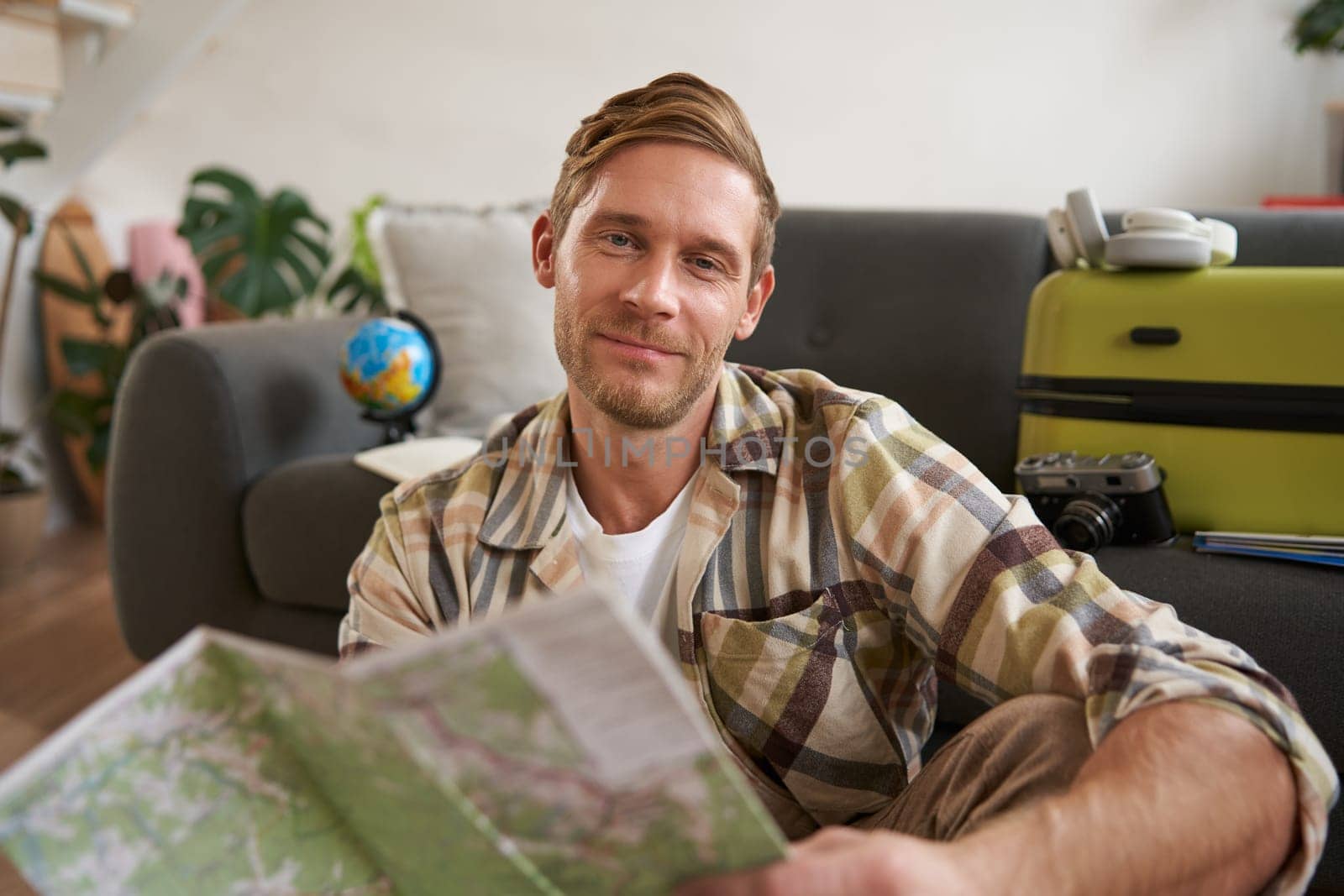 Close up portrait of smiling handsome man, boyfriend plans a holiday, holds travel map, sits near packed suitcase.