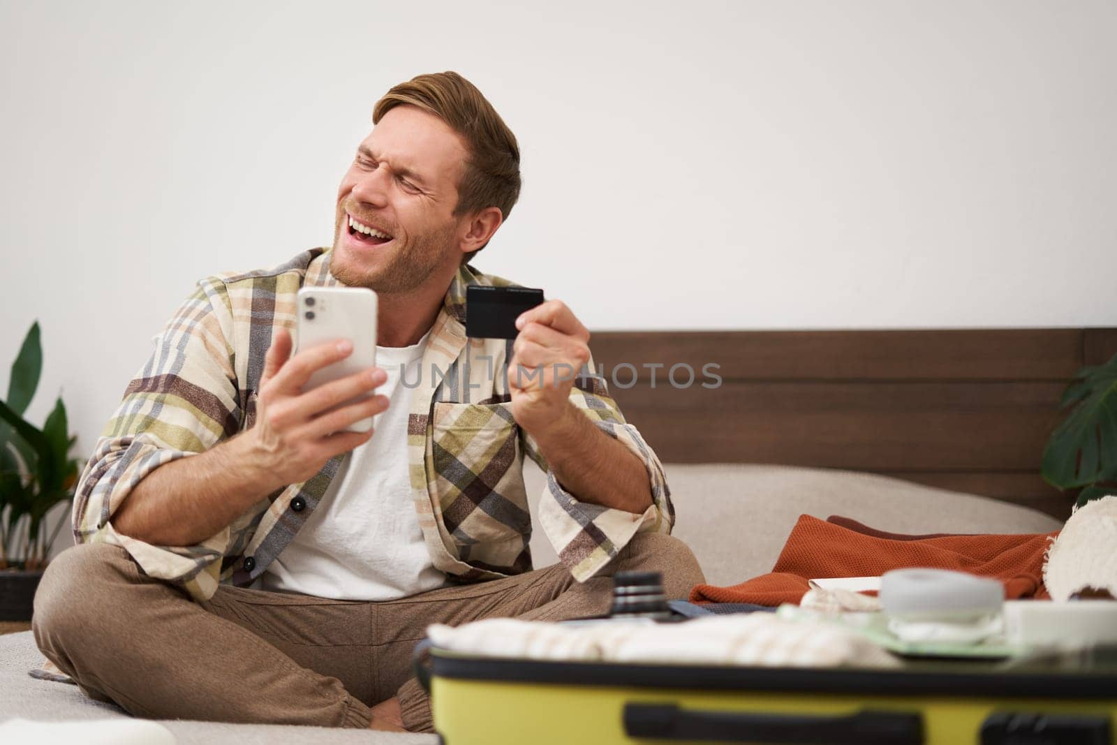 Cheerful tourist, guy with credit card and mobile phone, paying for holiday tickets, sitting on bed with suitcase, packing and going on holiday.