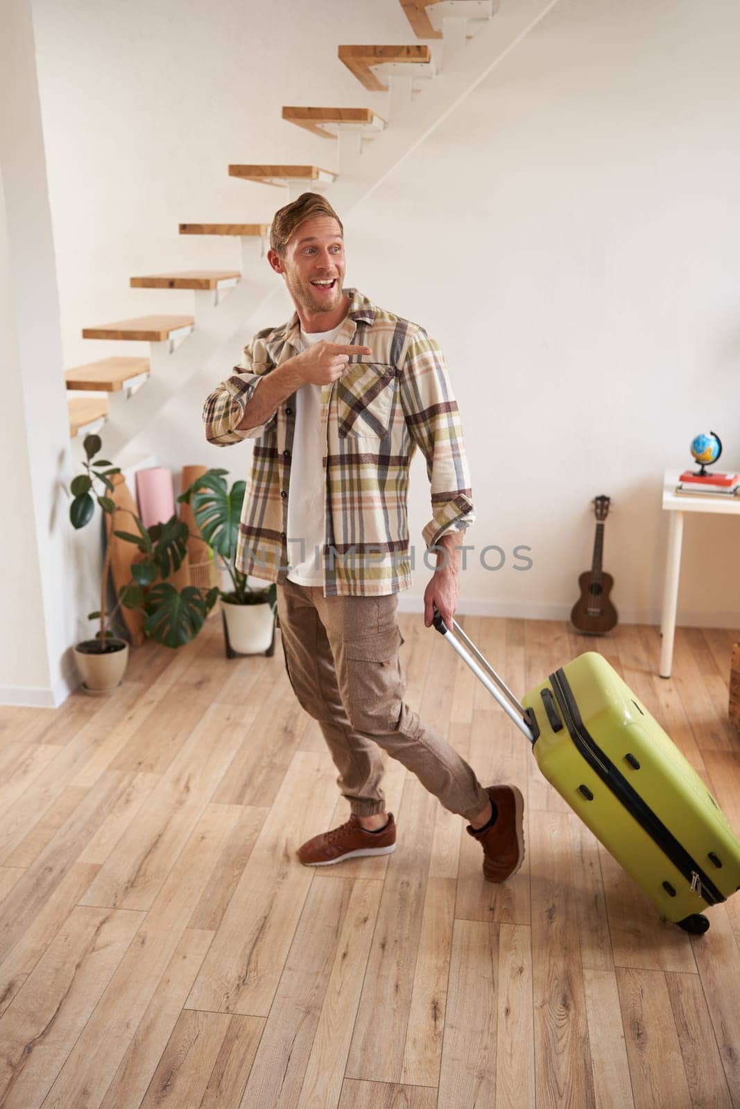 Full length portrait of handsome smiling man, pointing finger at someone, leaving the apartment with luggage, going on a trip, travelling by Benzoix