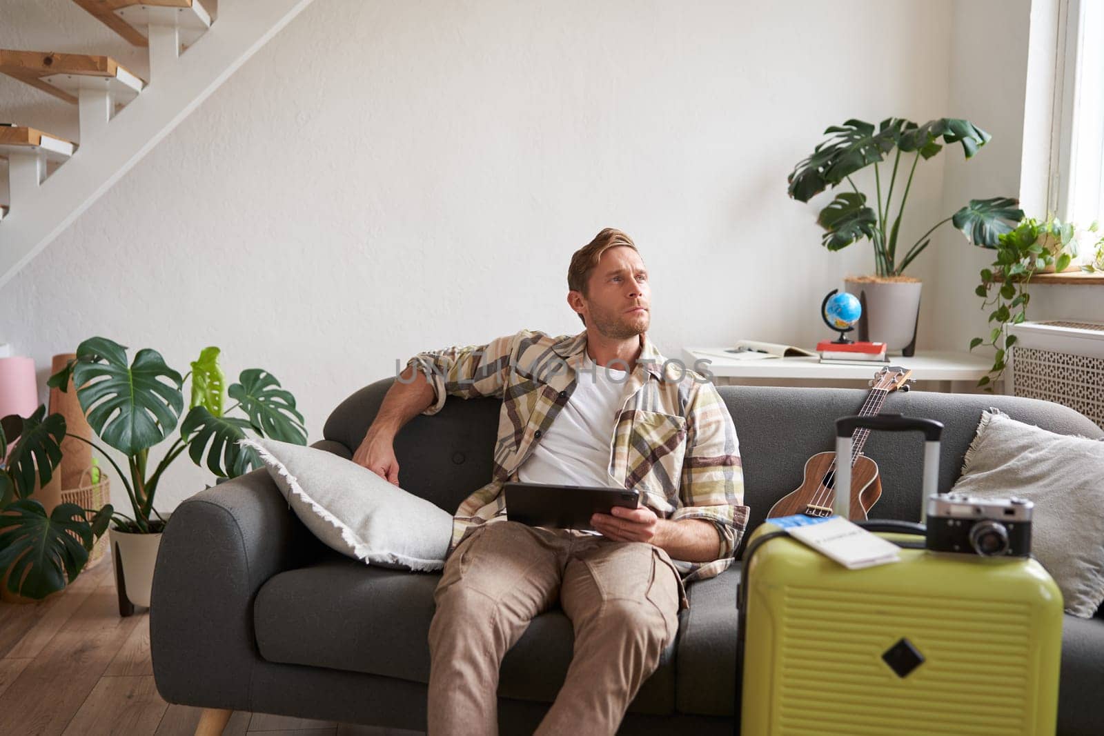 Image of young man with digital tablet, tourist sitting on sofa with suitcase packed and ukulele, thinking, looking aside thoughtful, planning holiday trip. Tourism concept