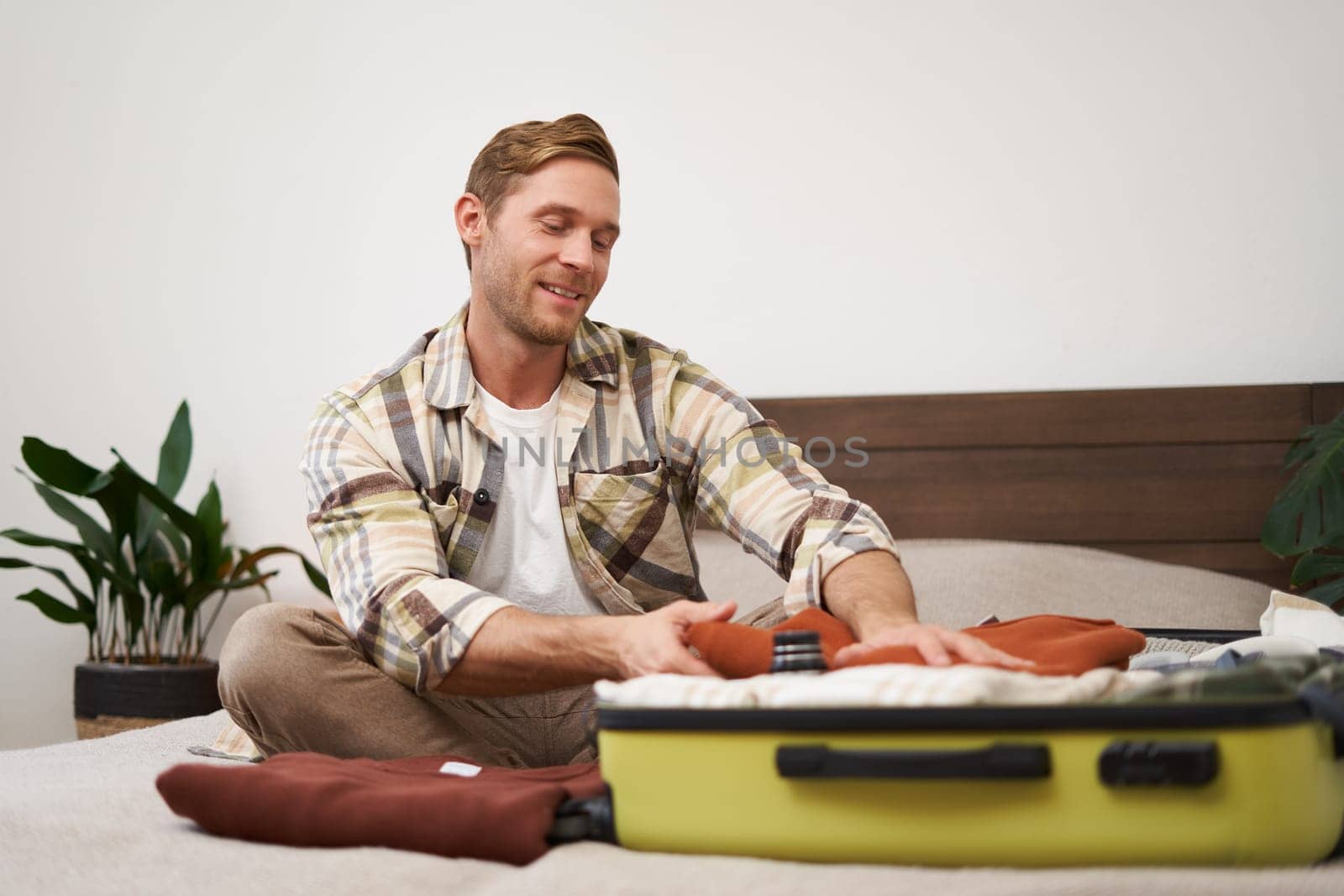 Portrait of smiling handsome man, sits on bed, puts clothes inside suitcase, goes on vacation, dreams about holiday, books a trip with travel agency and packs things. Lifestyle and tourism concept