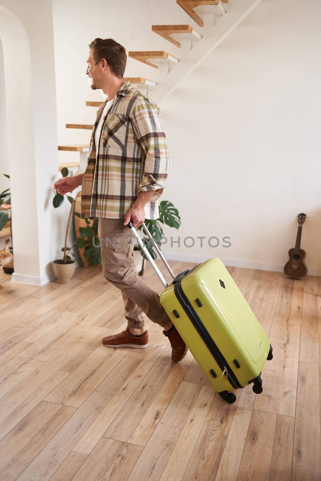 Vertical portrait of young handsome man walking indoors with suitcase, going on vacation, leaving his rented apartment with luggage.