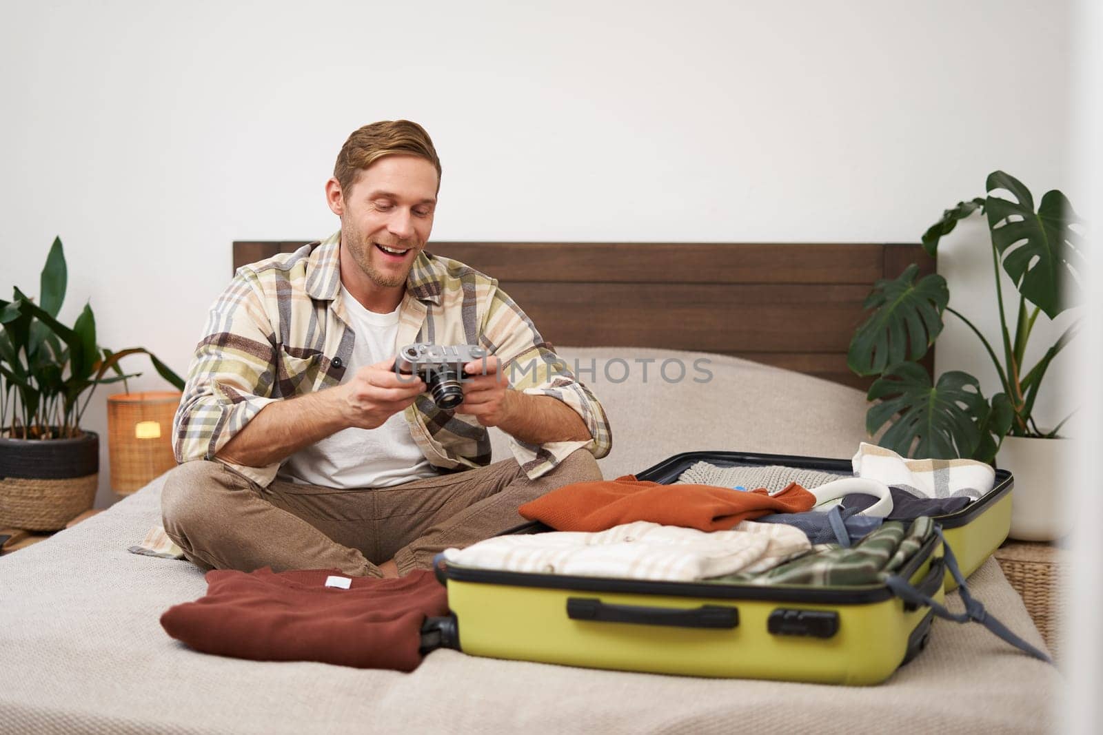 Portrait of handsome, happy young man, sitting with suitcase on bed, looking at his camera, laughing and smiling, watching photos or videos from holiday trip. Lifestyle concept