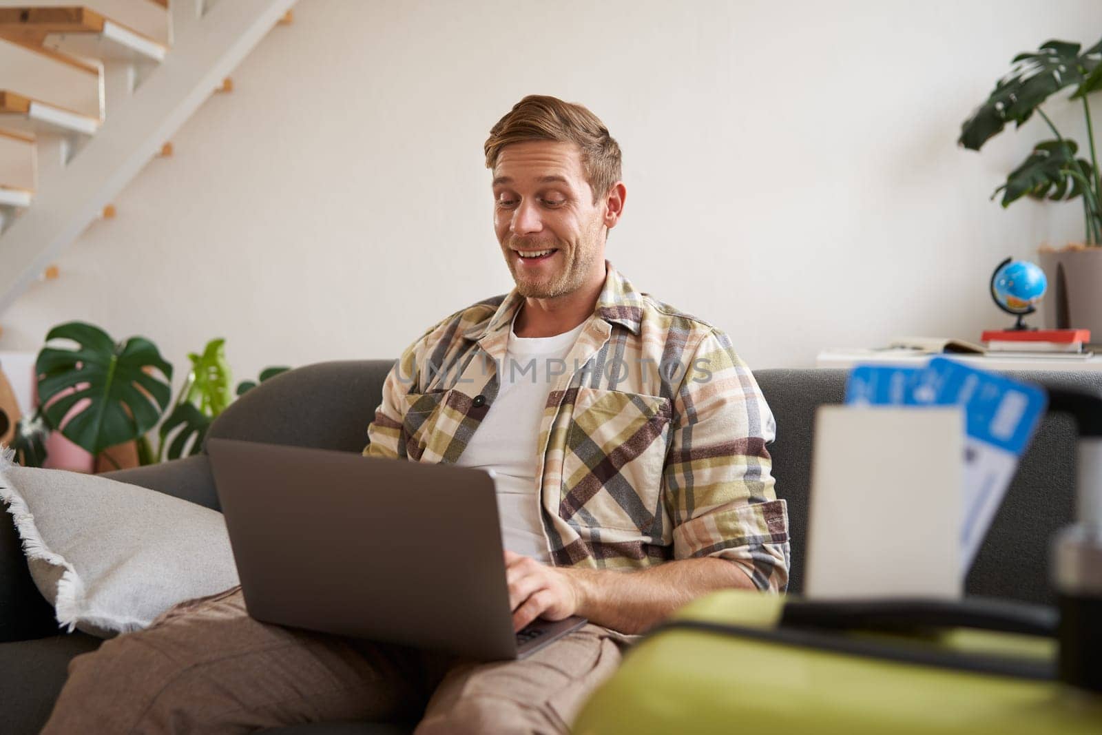 Portrait of happy young man booking holiday for two, has plane tickets, looks at laptop, sits in living room with computer by Benzoix