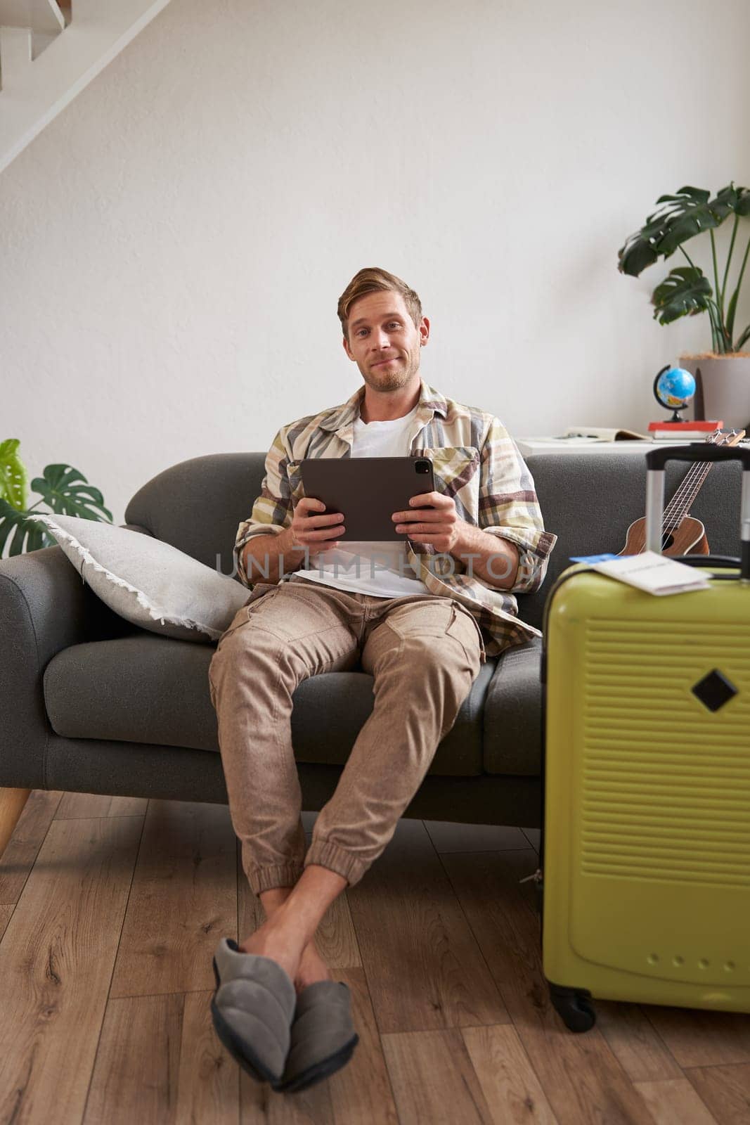 Vertical shot of young man going abroad, packing suitcase for a vacation, holding digital tablet, booking hotel online. Tourism and travel agency concept