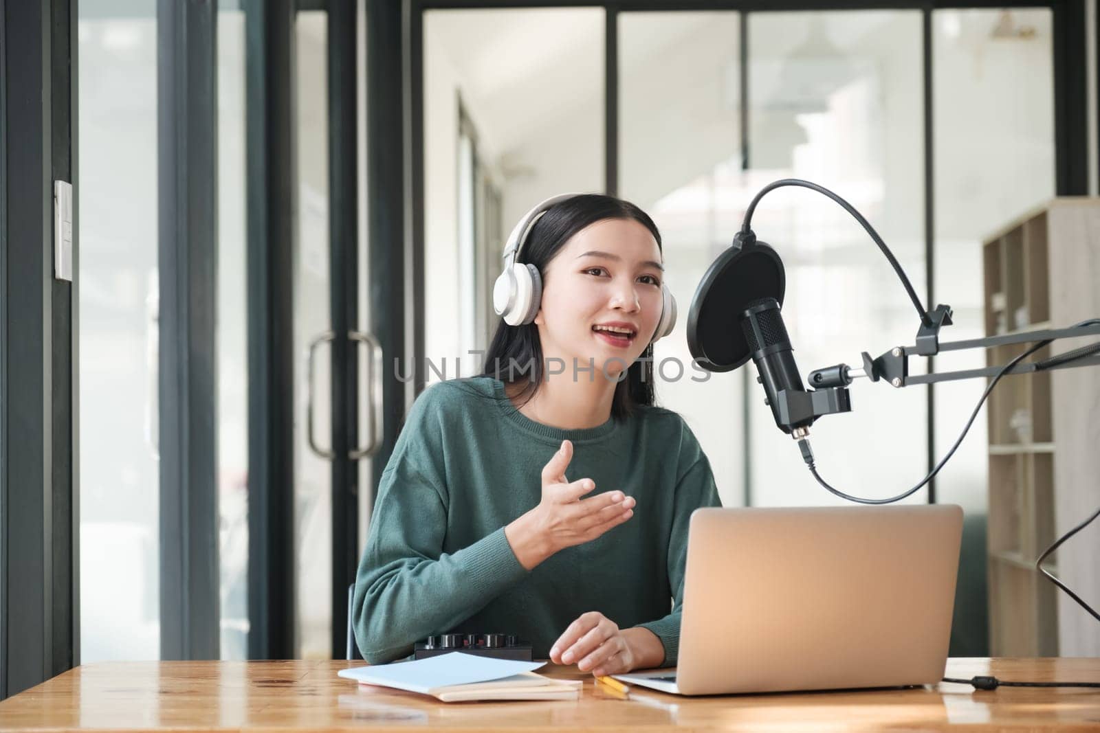 A woman is sitting at a desk with a laptop and a microphone. She is wearing headphones and she is talking into the microphone. The scene suggests that she might be a radio host or a podcast host