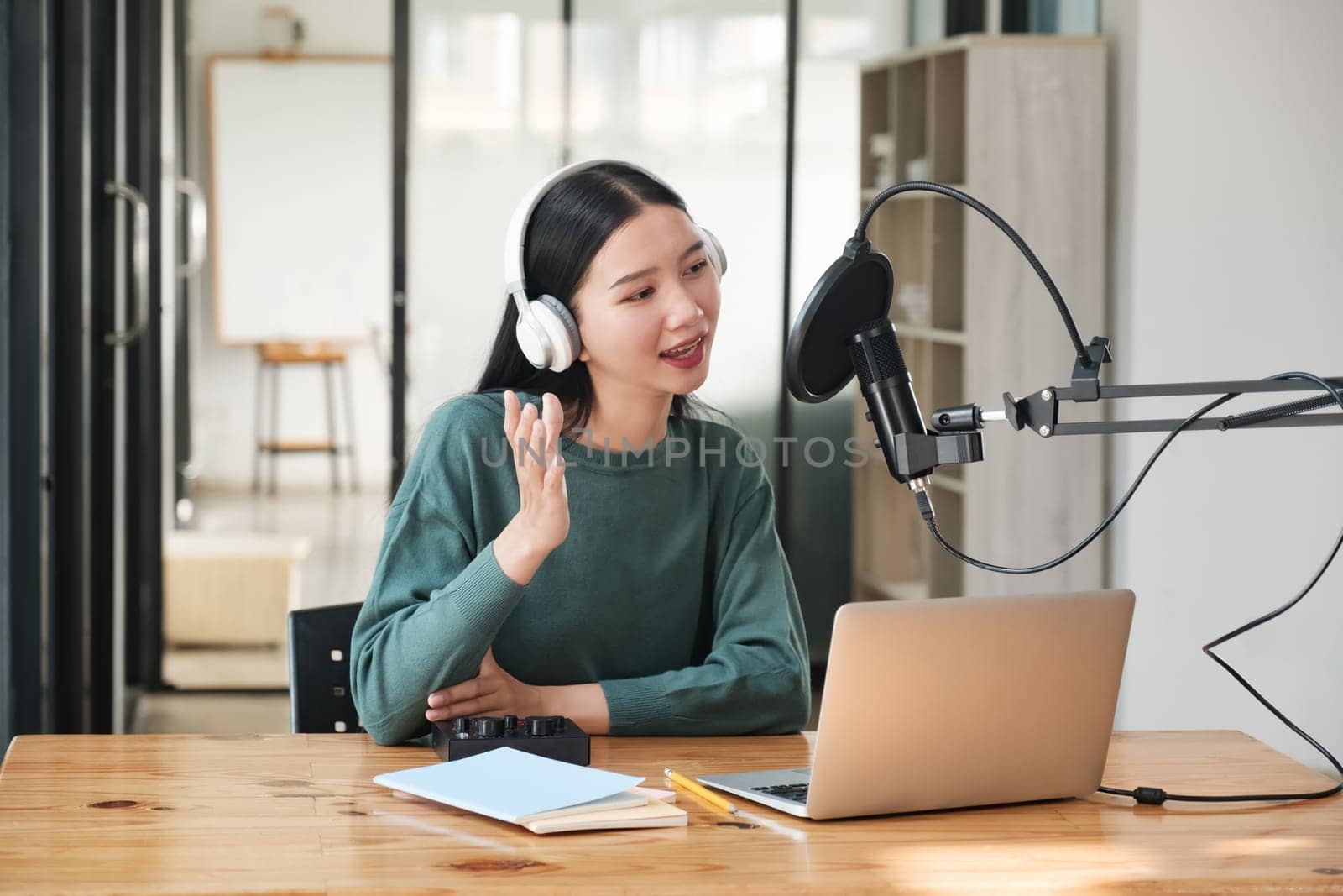 A woman wearing headphones is talking into a microphone. She is sitting at a desk with a laptop and a stack of papers. The scene suggests that she is working on a project or recording a podcast
