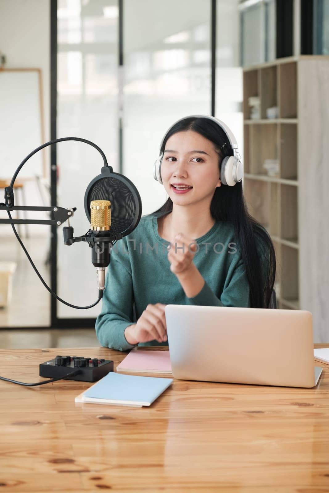 A woman is sitting at a desk with a microphone and a laptop. She is wearing headphones and she is recording a podcast. The room has a wooden floor and a few books on the desk