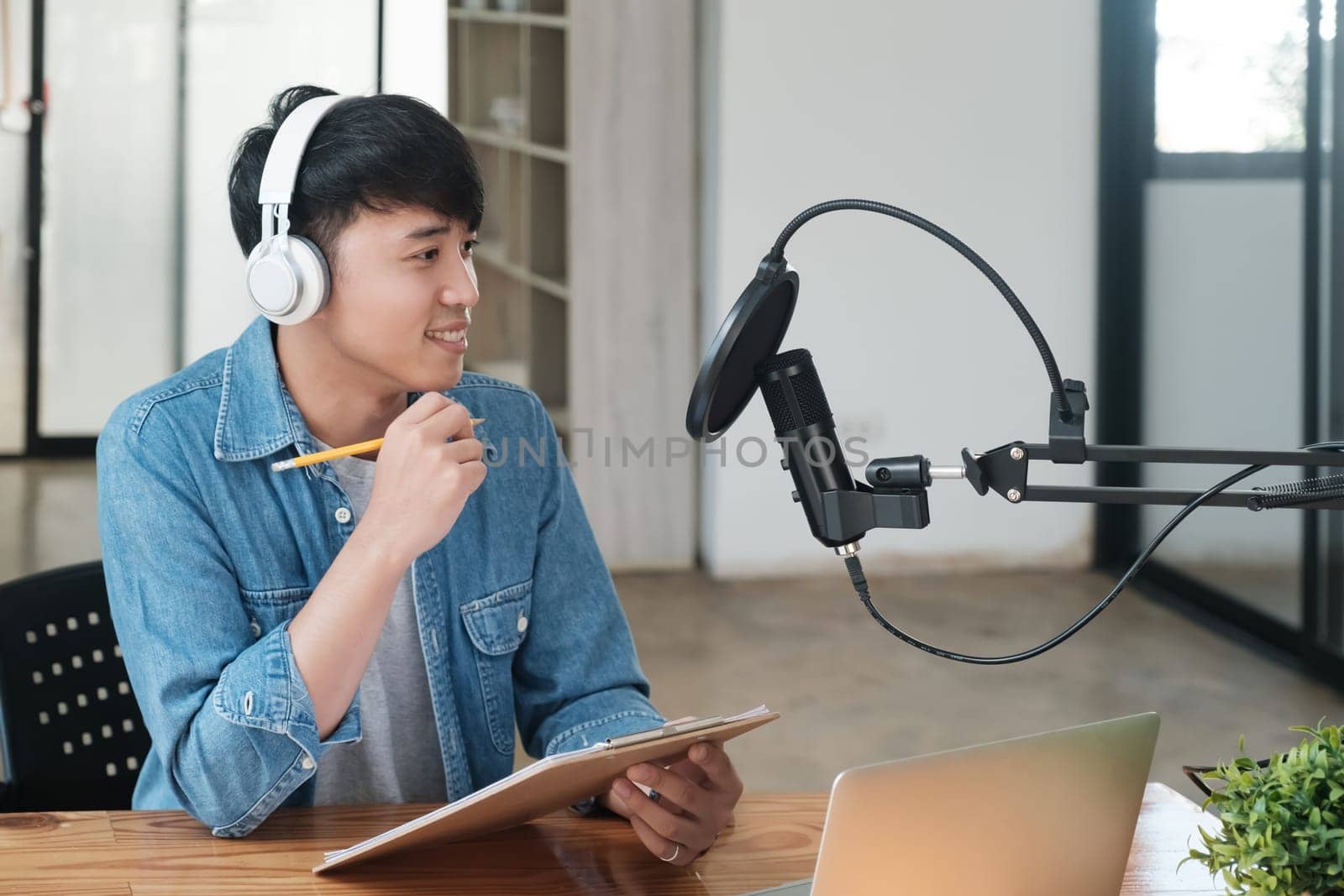 A man wearing headphones is sitting at a desk with a laptop and a notebook by ijeab