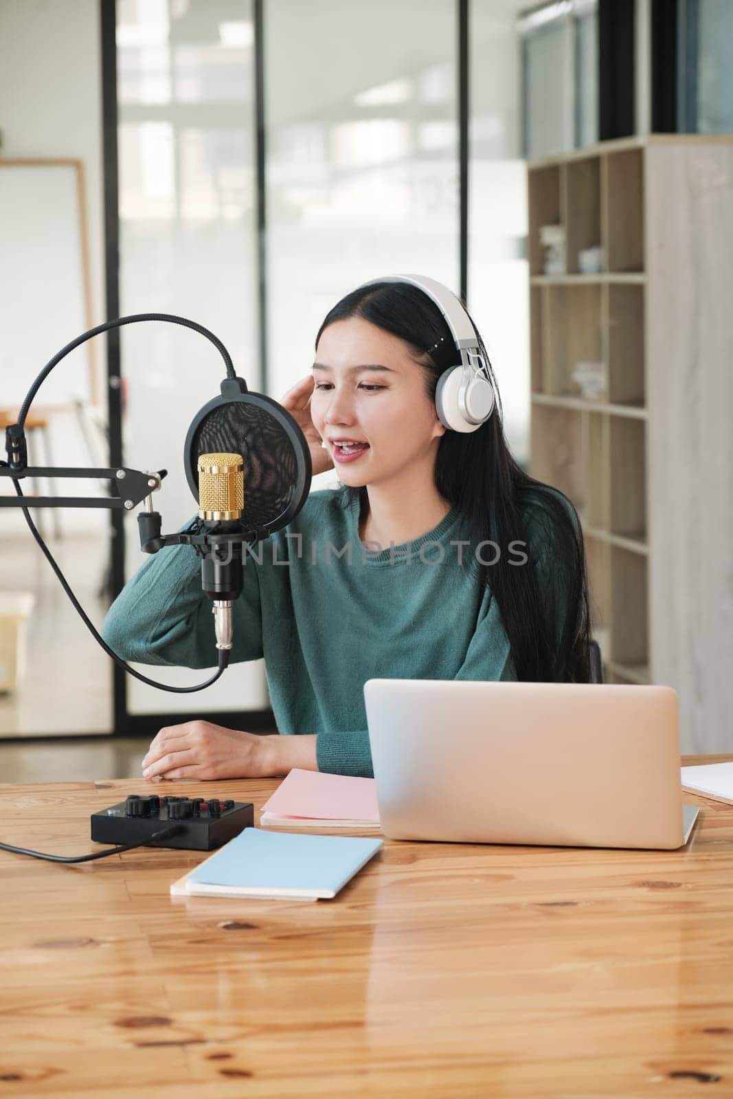 A woman is recording a song in a studio. She is wearing headphones and is sitting at a desk with a laptop. The room has a wooden table and a few books on it