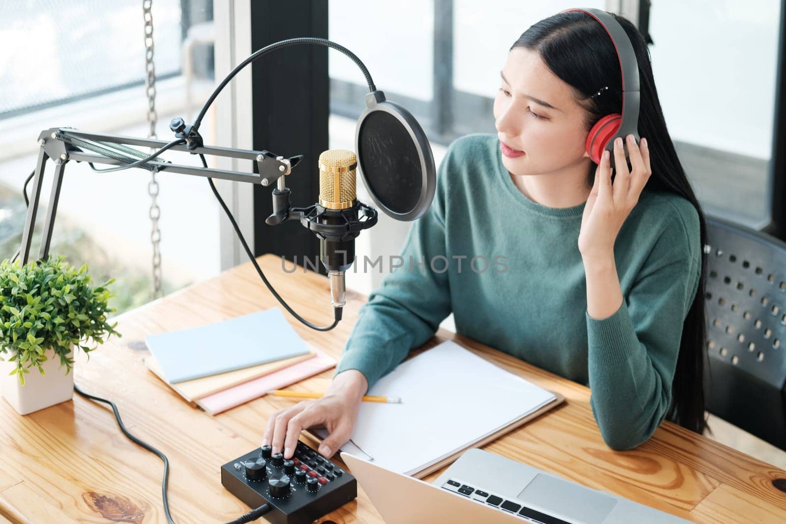 A woman is sitting at a desk with a microphone and a laptop. She is wearing headphones and she is recording a podcast or a voiceover