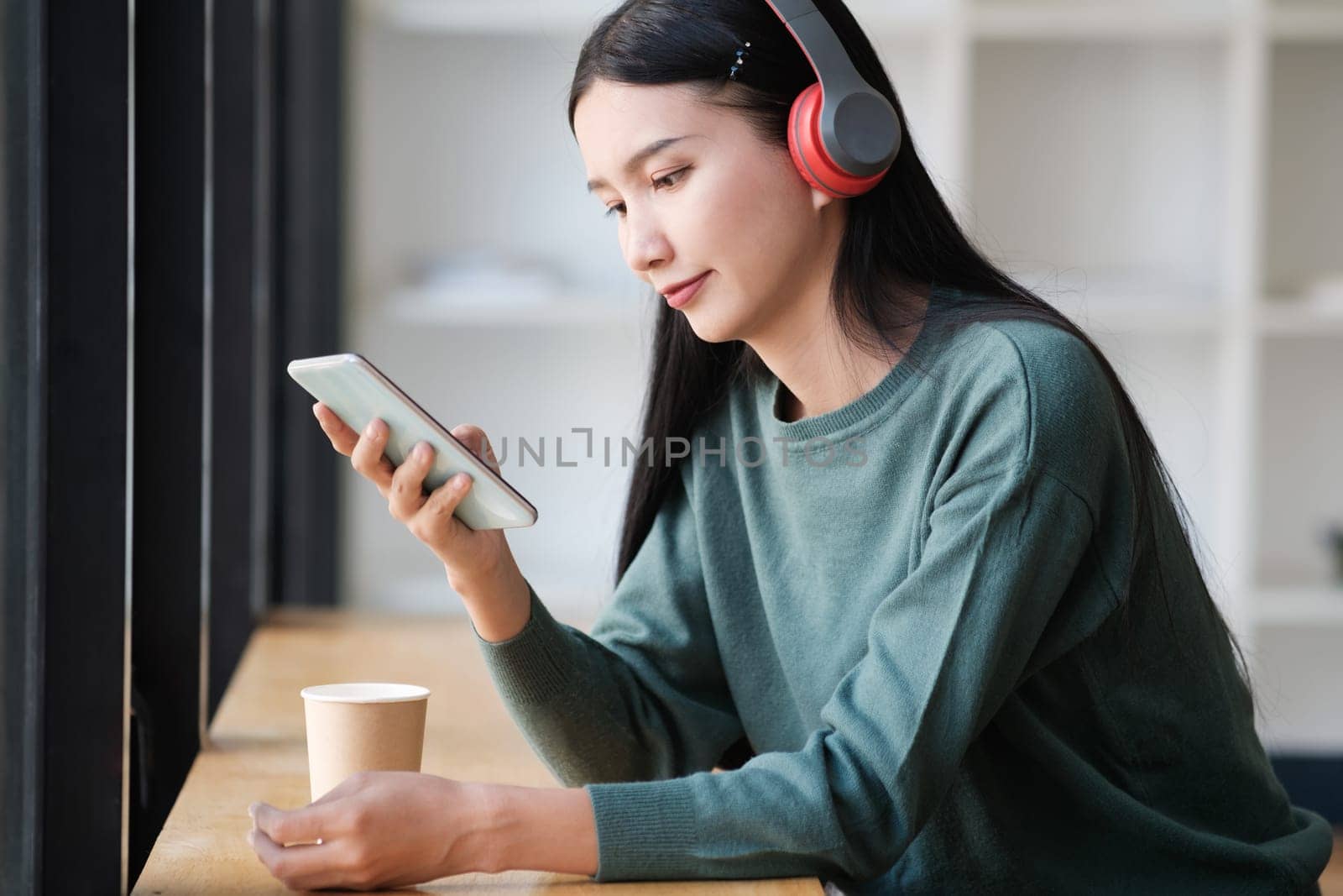 A woman is sitting at a table with a cell phone and a cup of coffee. She is wearing headphones and she is listening to music