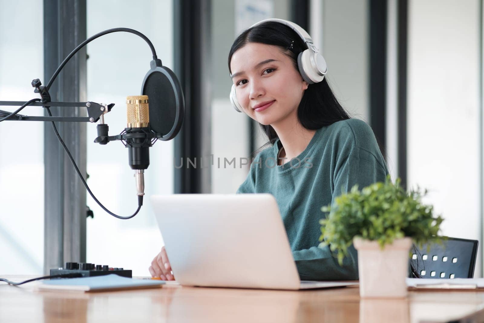 A woman is sitting at a desk with a laptop and a microphone. She is wearing headphones and smiling. The scene suggests that she is working on a project or recording something