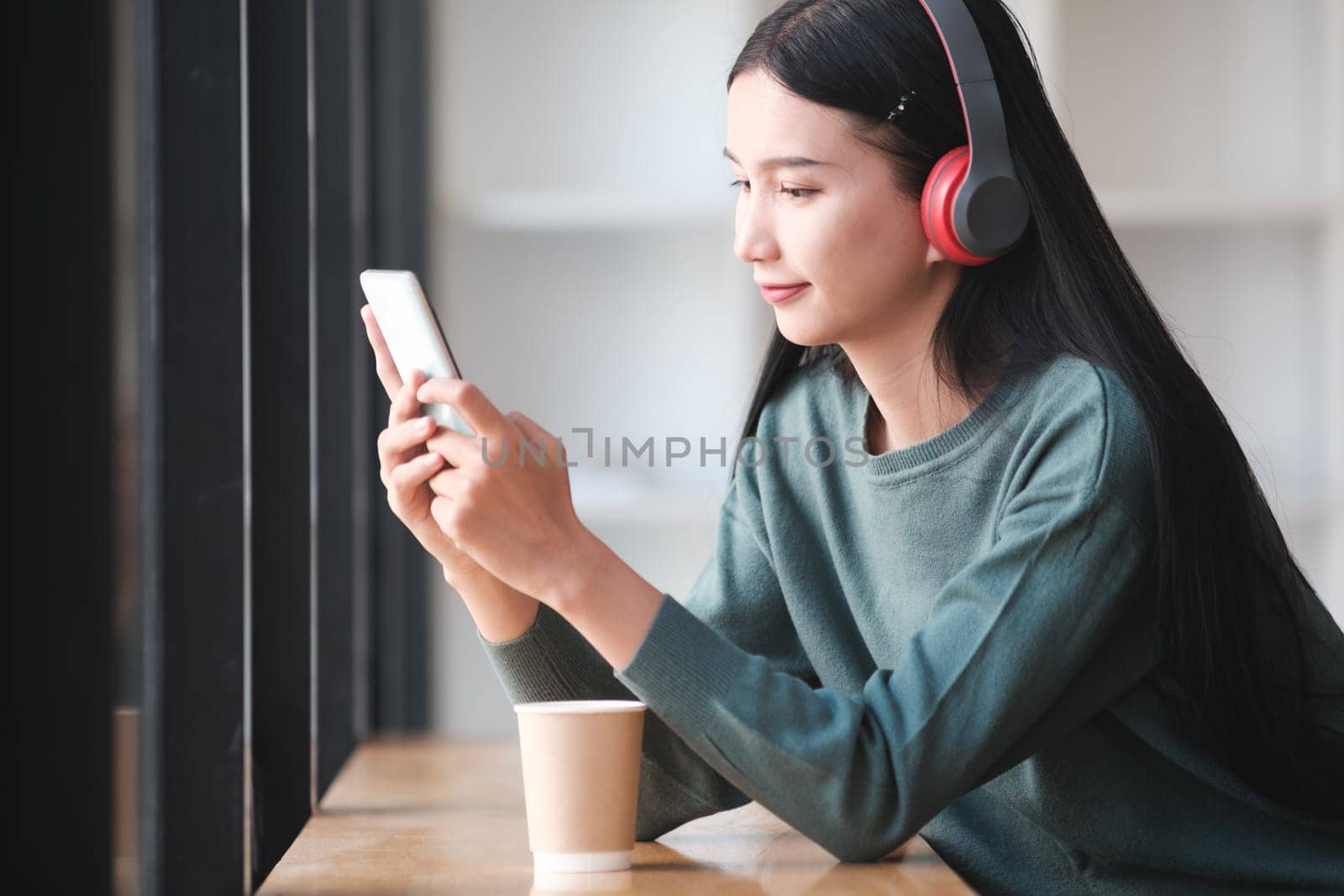 A woman is sitting at a table with a cup of coffee and a cell phone. She is wearing headphones and she is listening to music