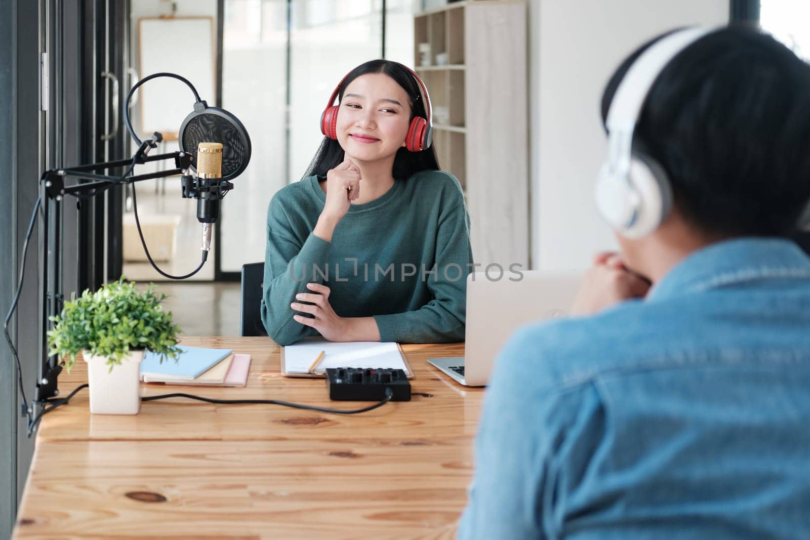 A woman is sitting at a desk with a microphone and a laptop. She is wearing headphones and smiling. The scene suggests a recording session or a podcast being produced