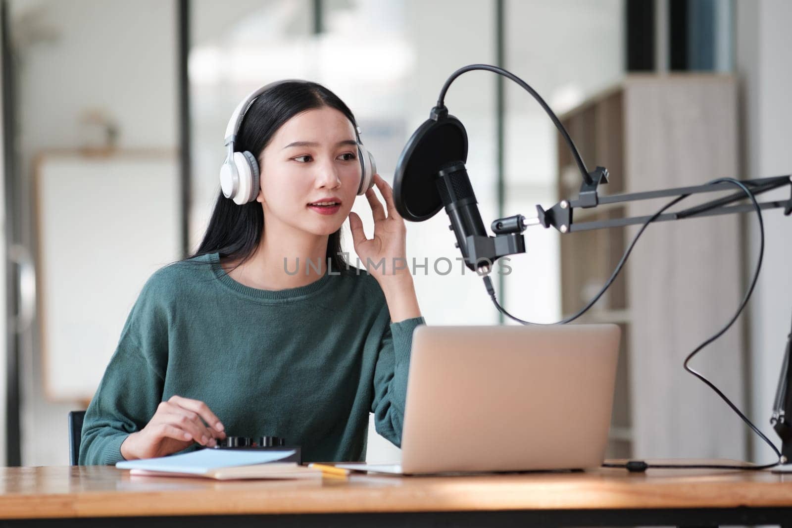 A woman wearing headphones is sitting at a desk with a laptop and a microphone by ijeab