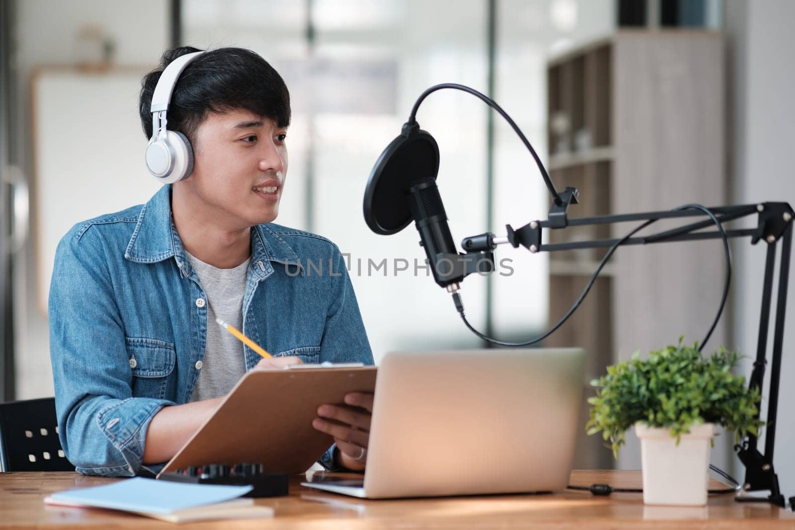 A man wearing headphones is sitting at a desk with a laptop and a clipboard by ijeab