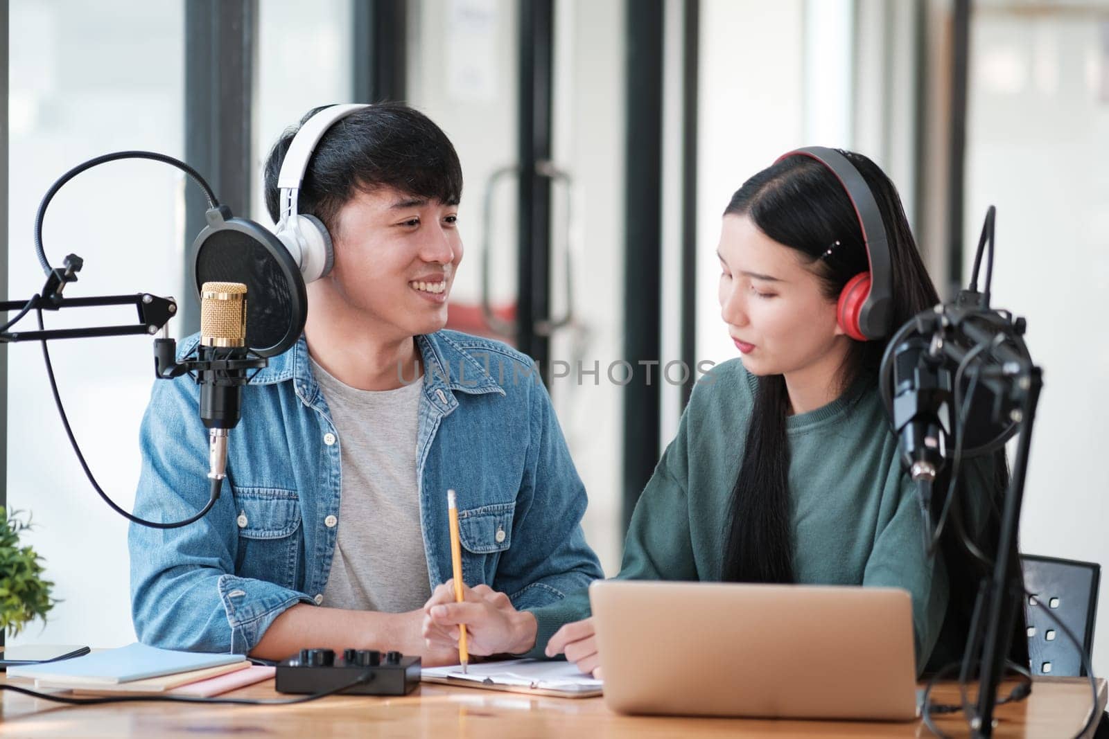 Two people are sitting at a desk with a laptop and a microphone. They are smiling and seem to be enjoying each other's company