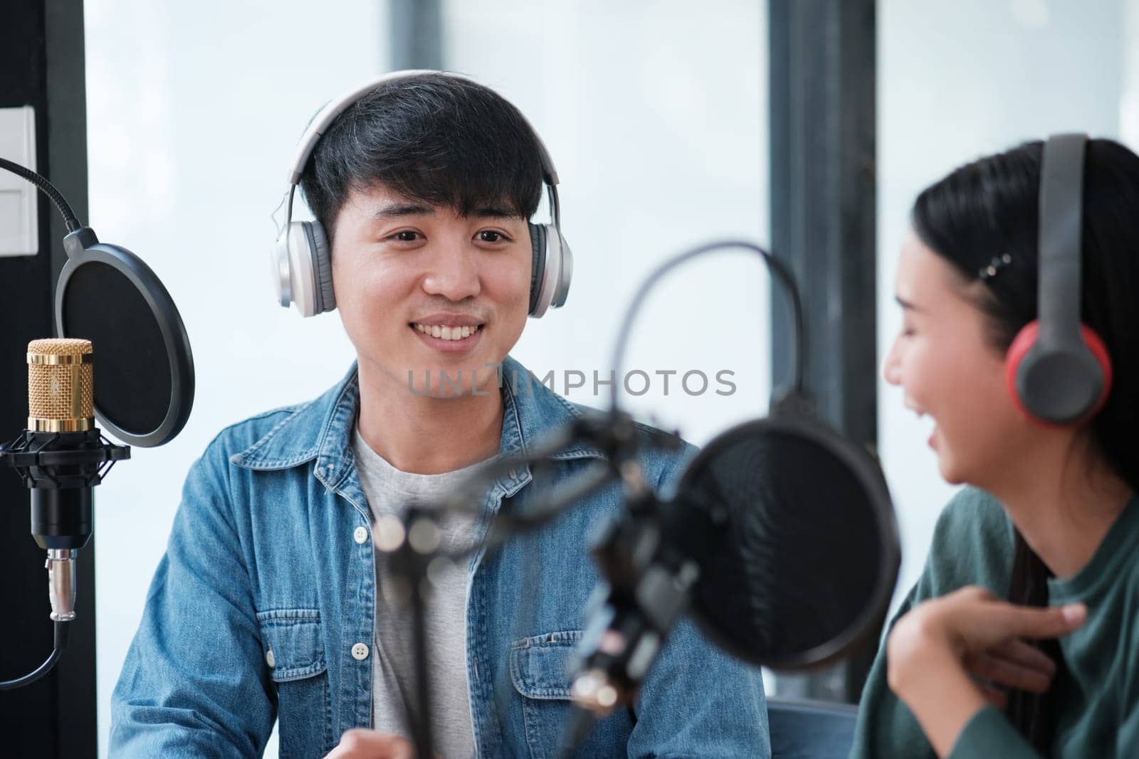 Two people are sitting in a studio with microphones and headphones on. One of them is smiling and the other is laughing. Scene is lighthearted and fun