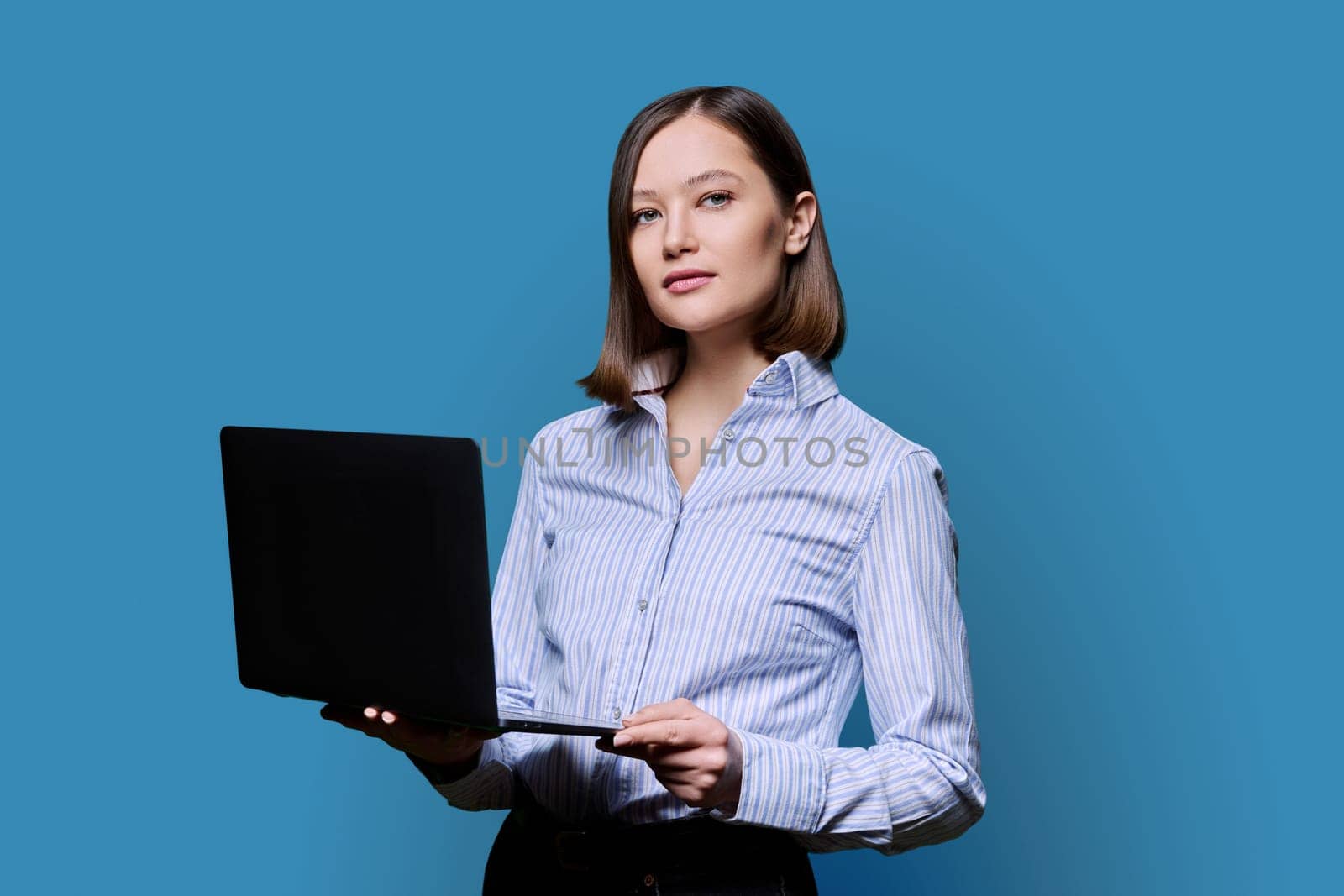 Serious young business woman with laptop on blue background. Confident successful female holding computer in hands looking at camera. Business, work, training, education, modern digital technologies