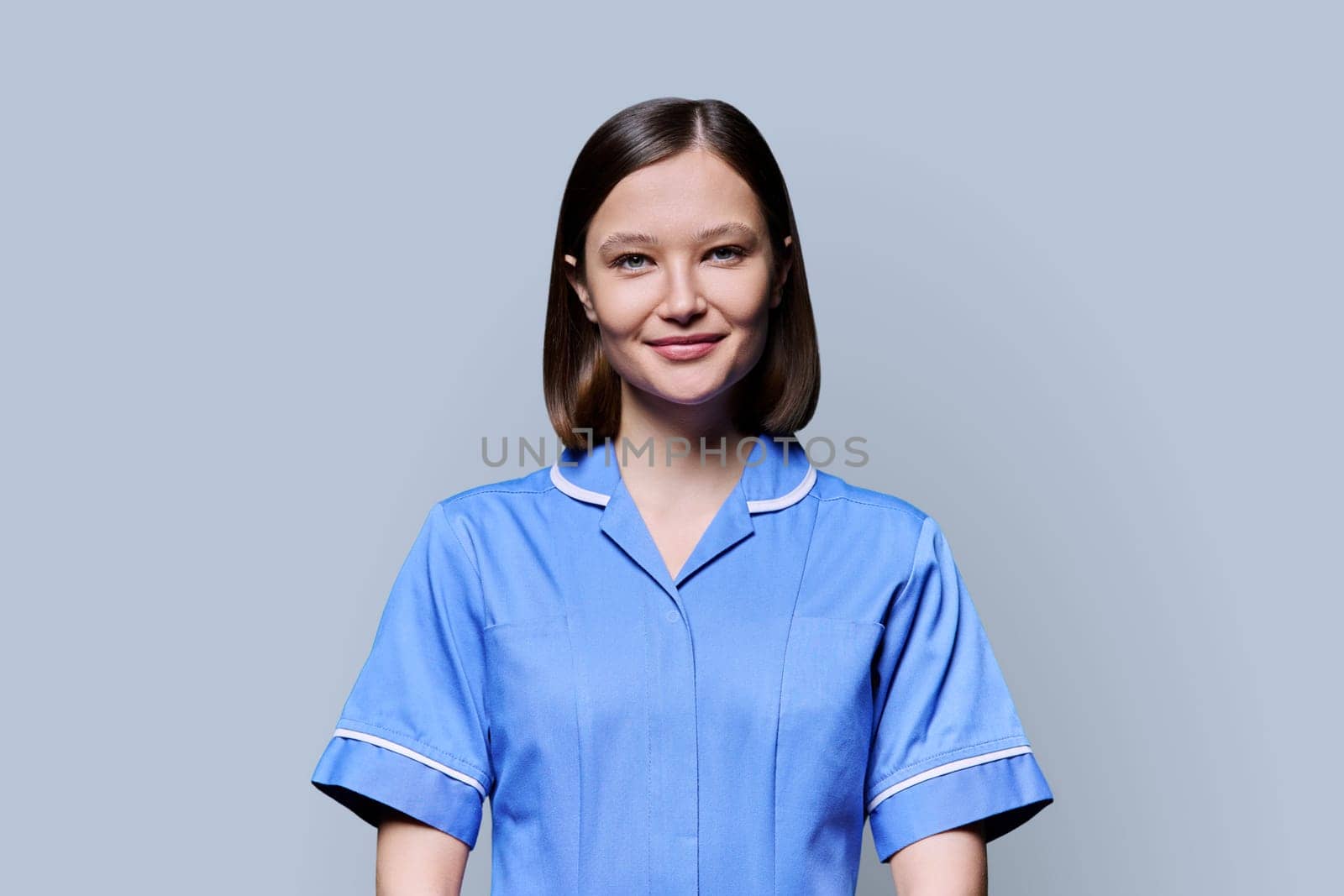 Portrait of young confident smiling female nurse looking at camera on gray studio background. Medical services, health, professional assistance, medical care concept